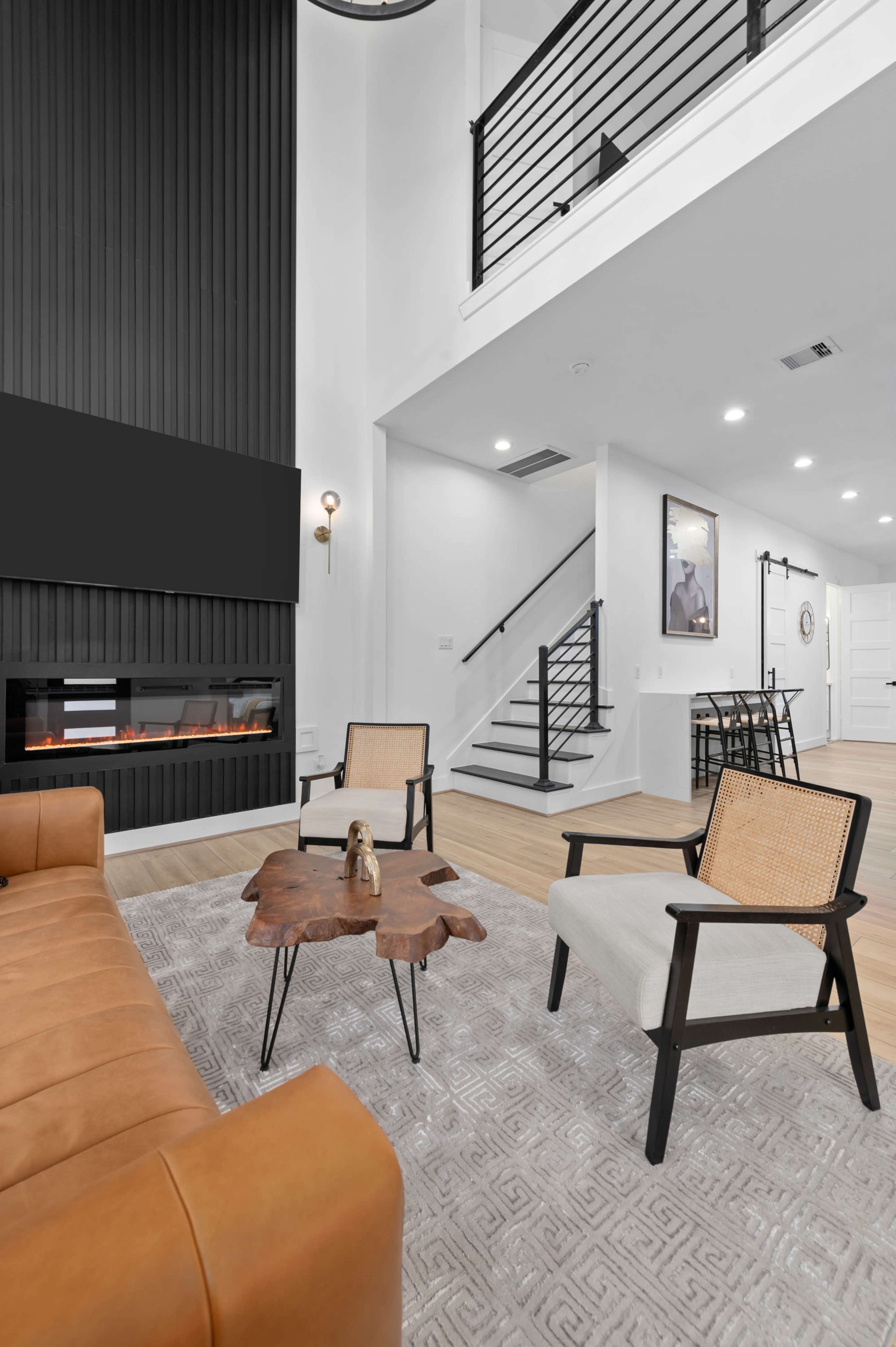 A modern living room features a tan leather sofa, two light-colored chairs, and a wooden coffee table on a patterned rug, with a staircase and a black-framed wall-mounted television in the background.