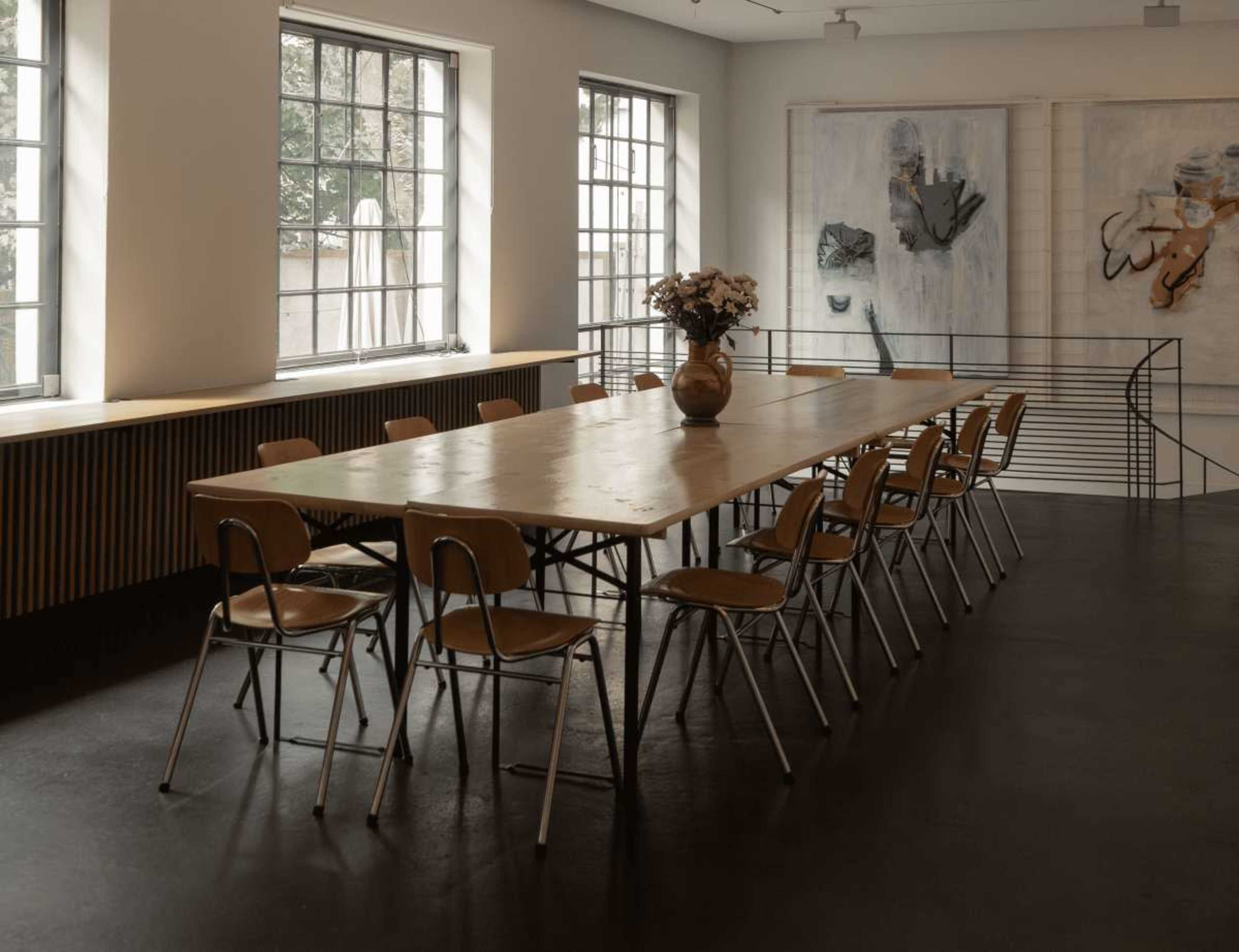 A long wooden table with twelve metal-legged chairs is set in a brightly lit room featuring large windows and abstract paintings on the walls.