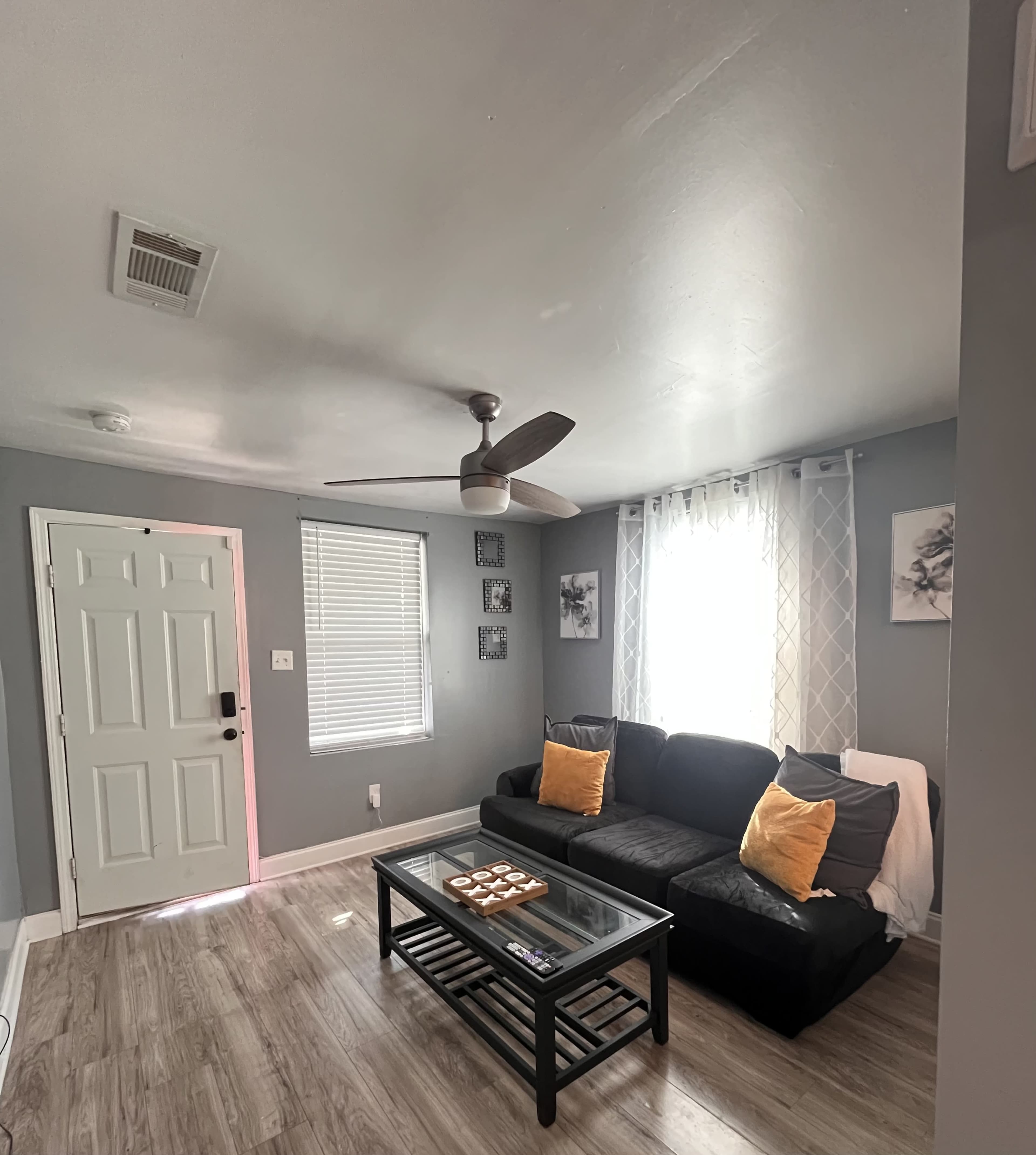 The image shows a living room with a black sofa, a glass coffee table, and light-colored walls, featuring a door and windows with blinds.
