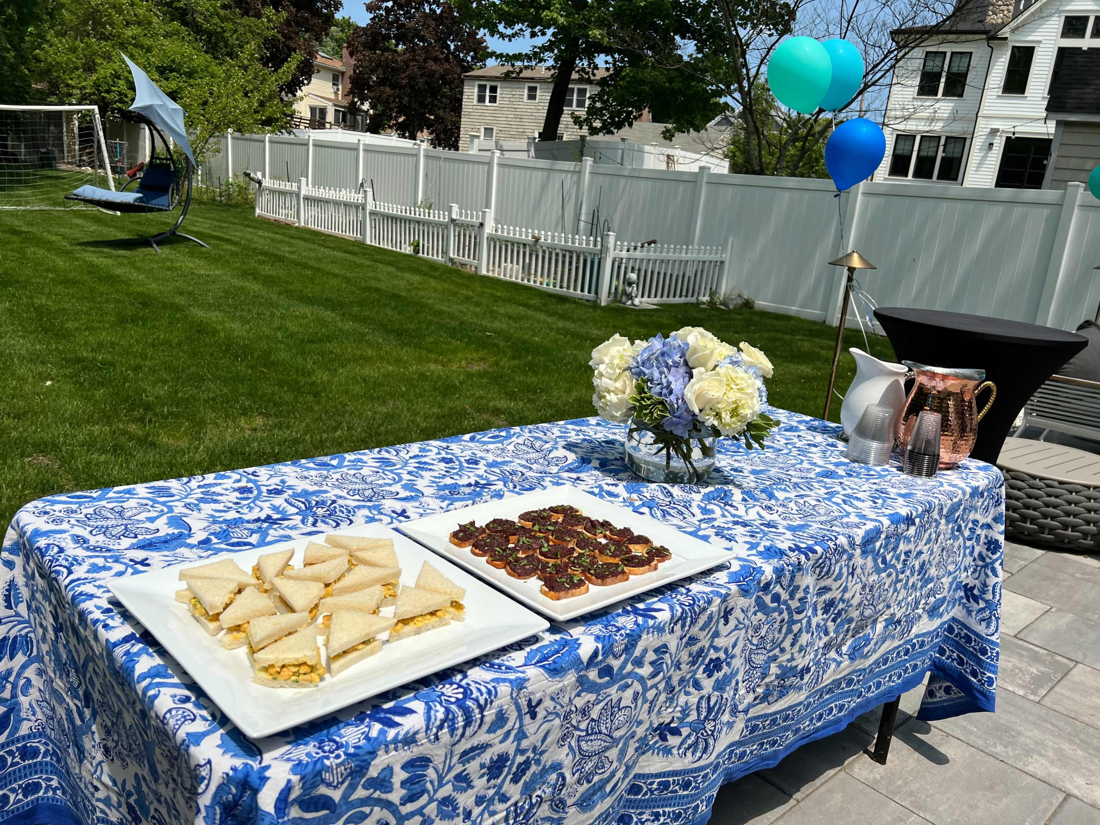 A table covered with a blue and white patterned tablecloth displays various food items, including triangular pastries and a tray of desserts, set in a grassy backyard with decorative balloons in the background.