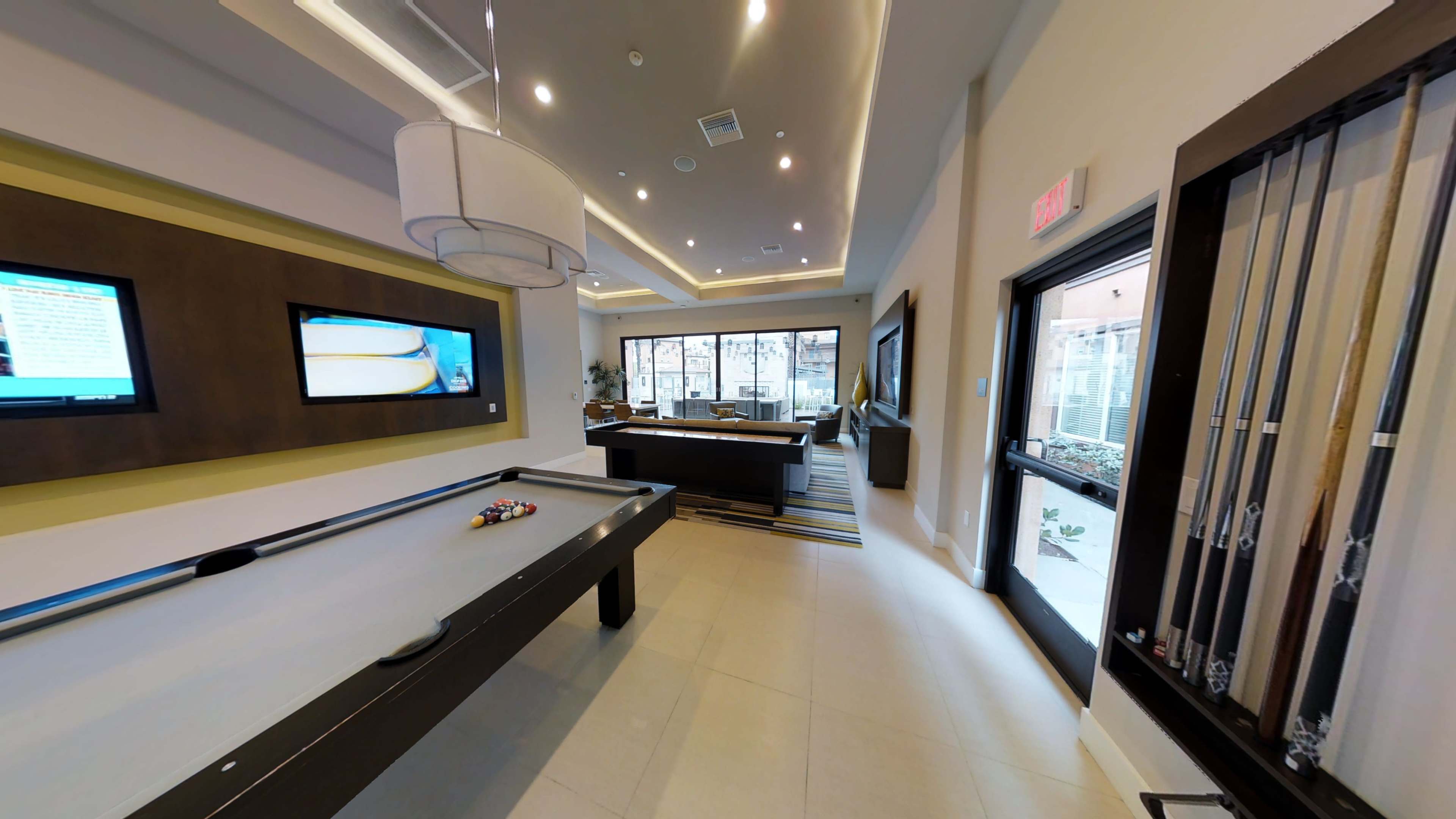 A modern recreational room with a billiard table, televisions mounted on the wall, and large windows overlooking an outdoor area.