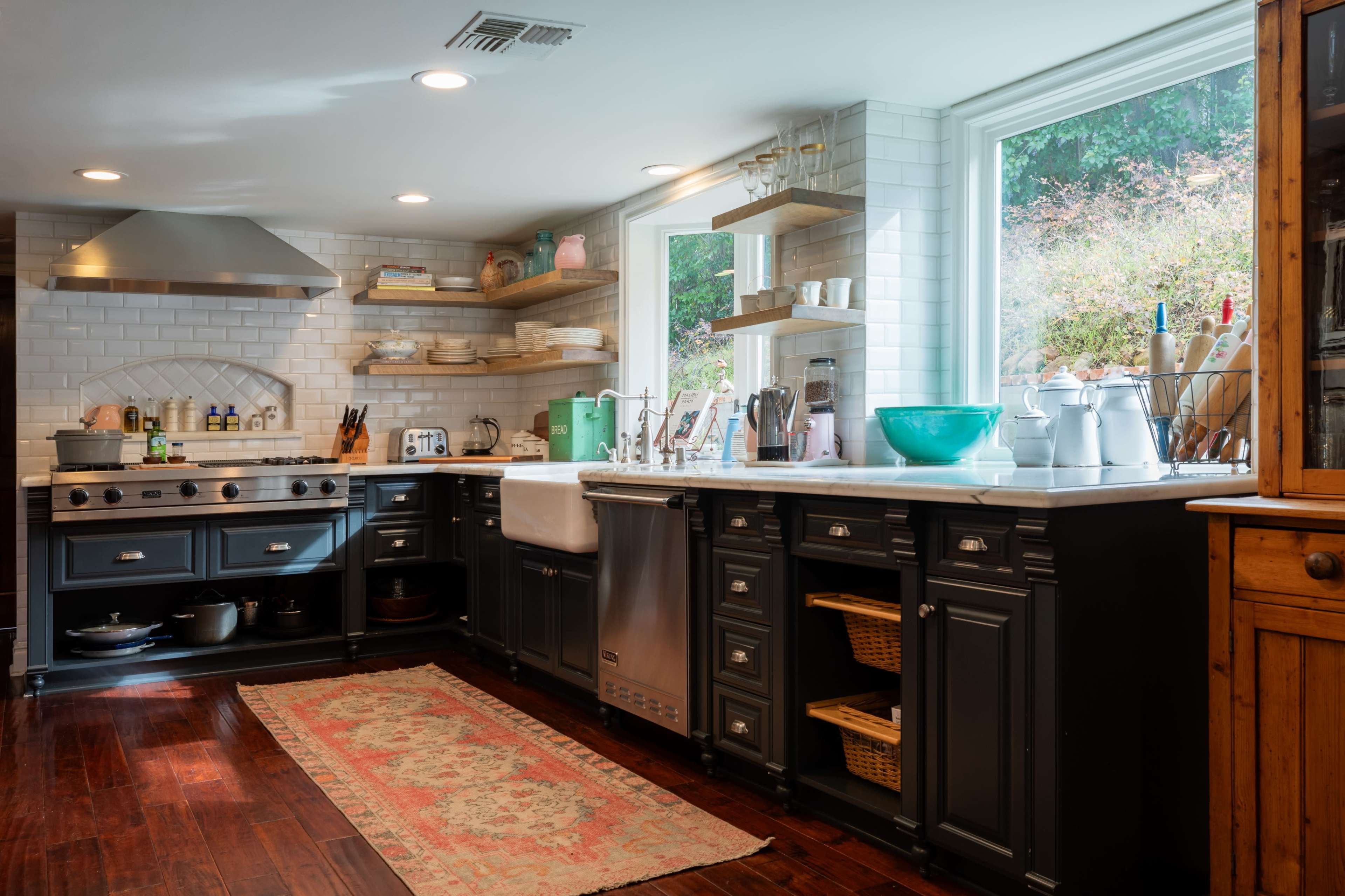 A modern kitchen features dark cabinetry, a large farmhouse sink, stainless steel appliances, open shelves with dishware, and a patterned rug on hardwood flooring.