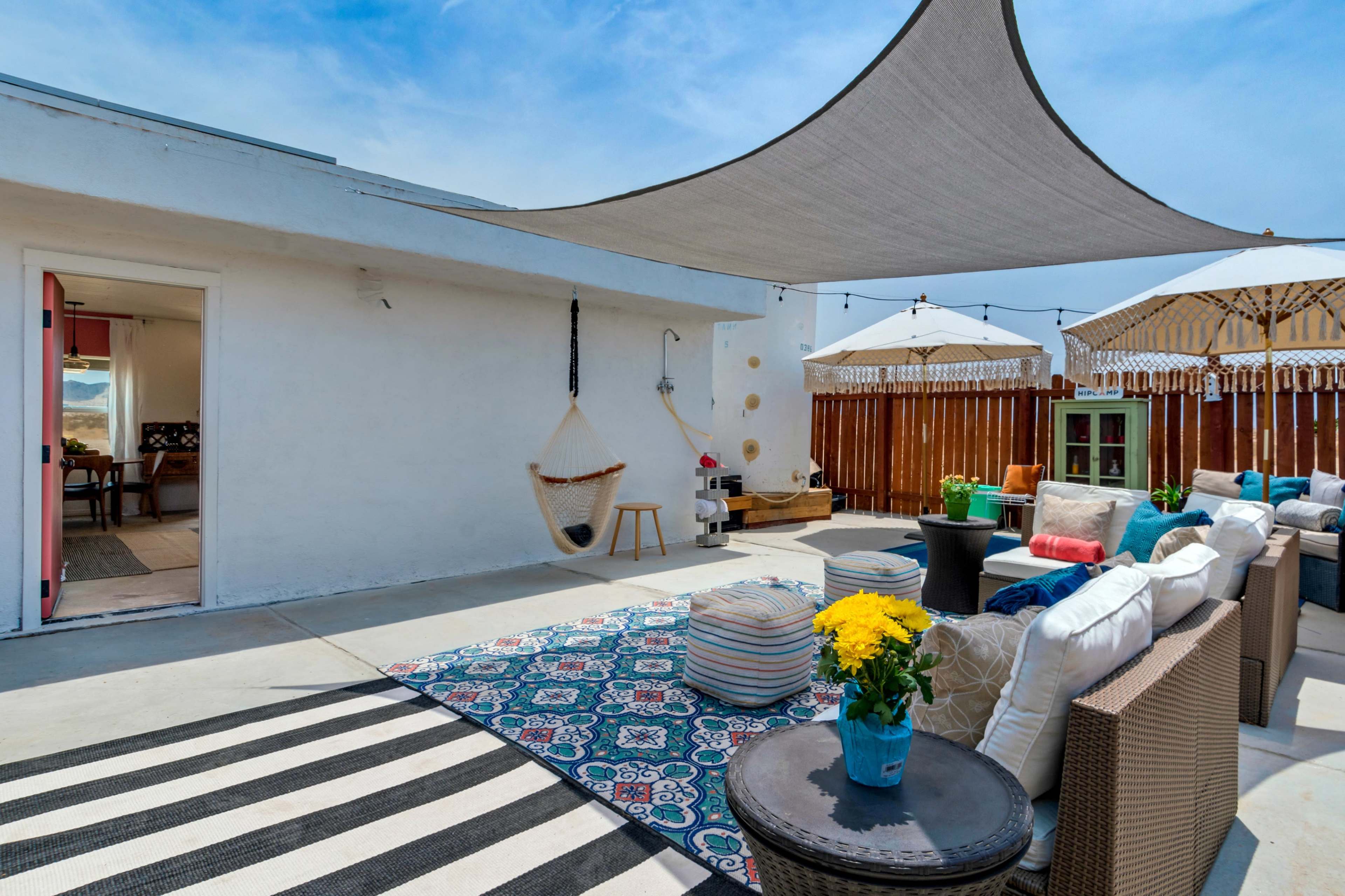 The image depicts a patio area with a striped rug, lounge seating, a hanging chair, and umbrellas, surrounded by a wooden fence.