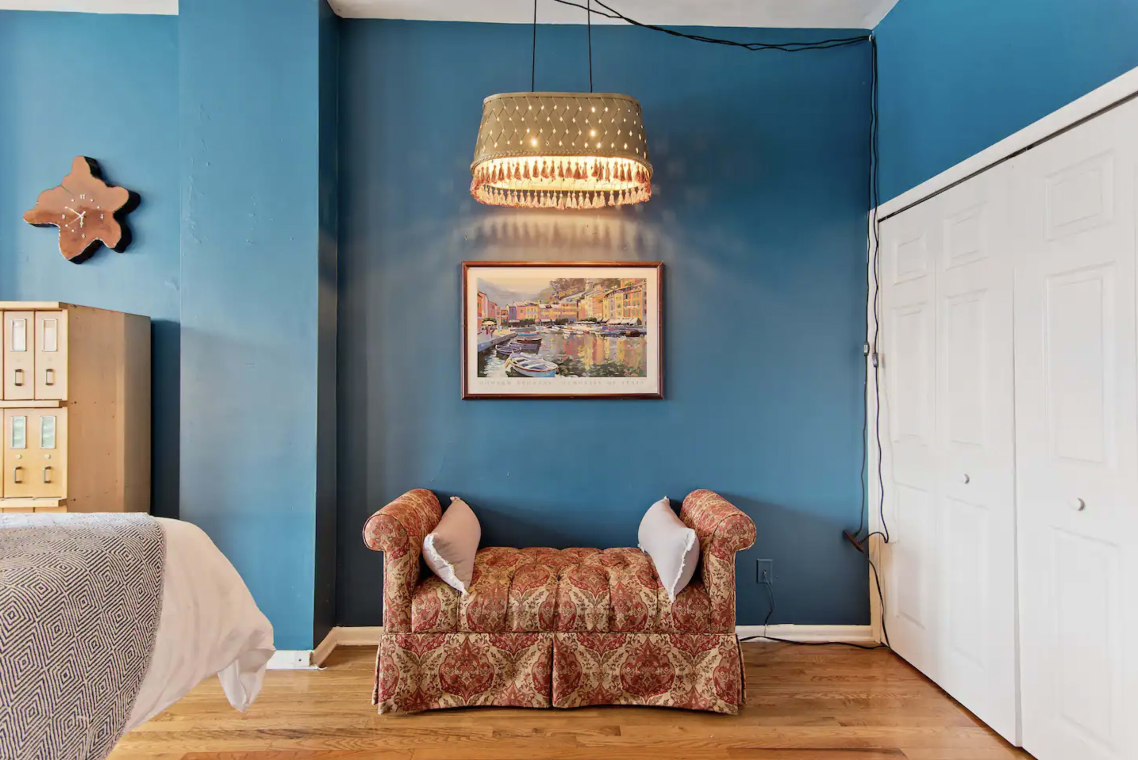 A room with a blue wall, a patterned loveseat with cushions, a framed painting, and a pendant light hanging above.