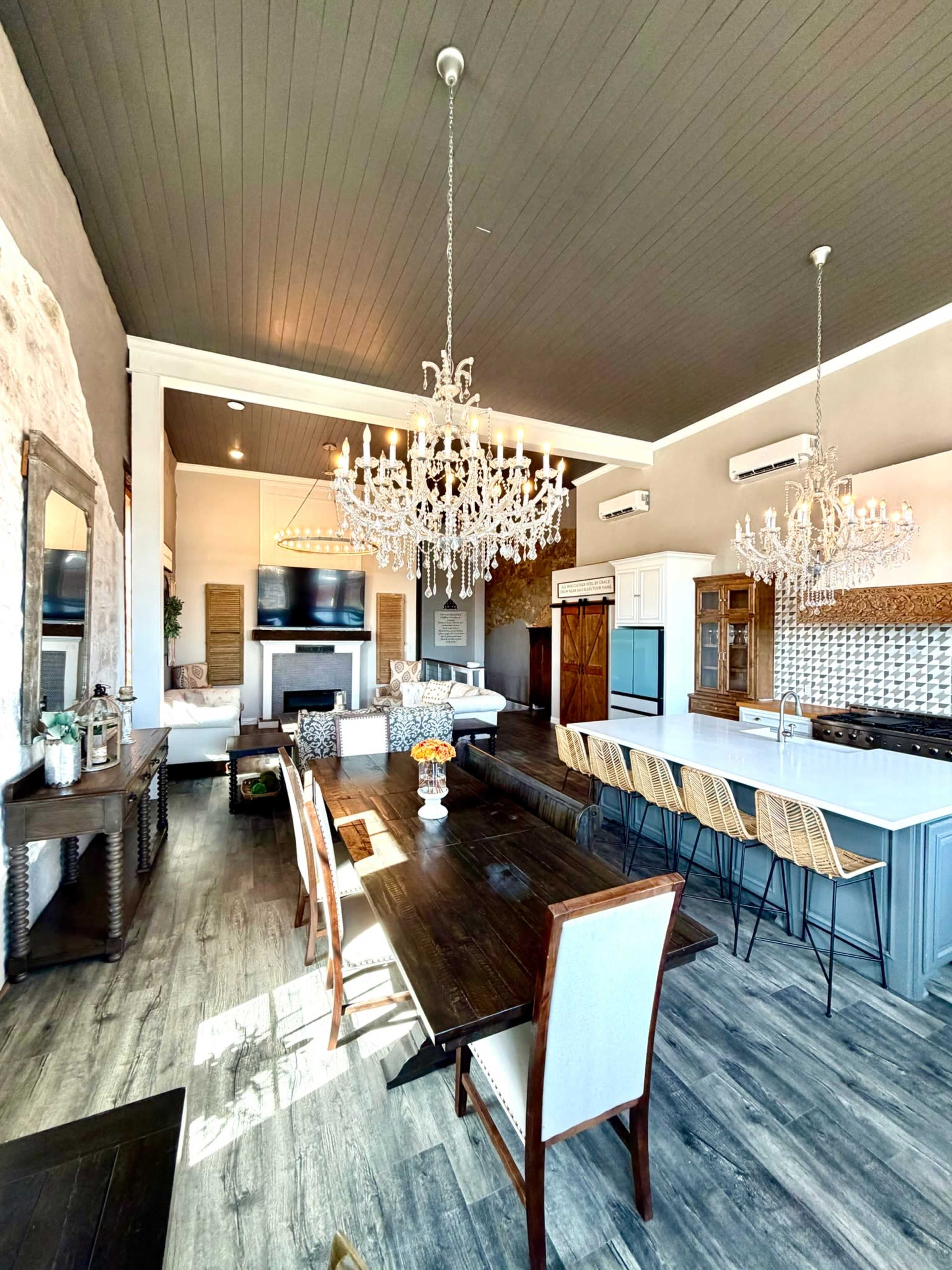 The image depicts a spacious kitchen and dining area featuring a large wooden table, elegant chandeliers, and modern furnishings.