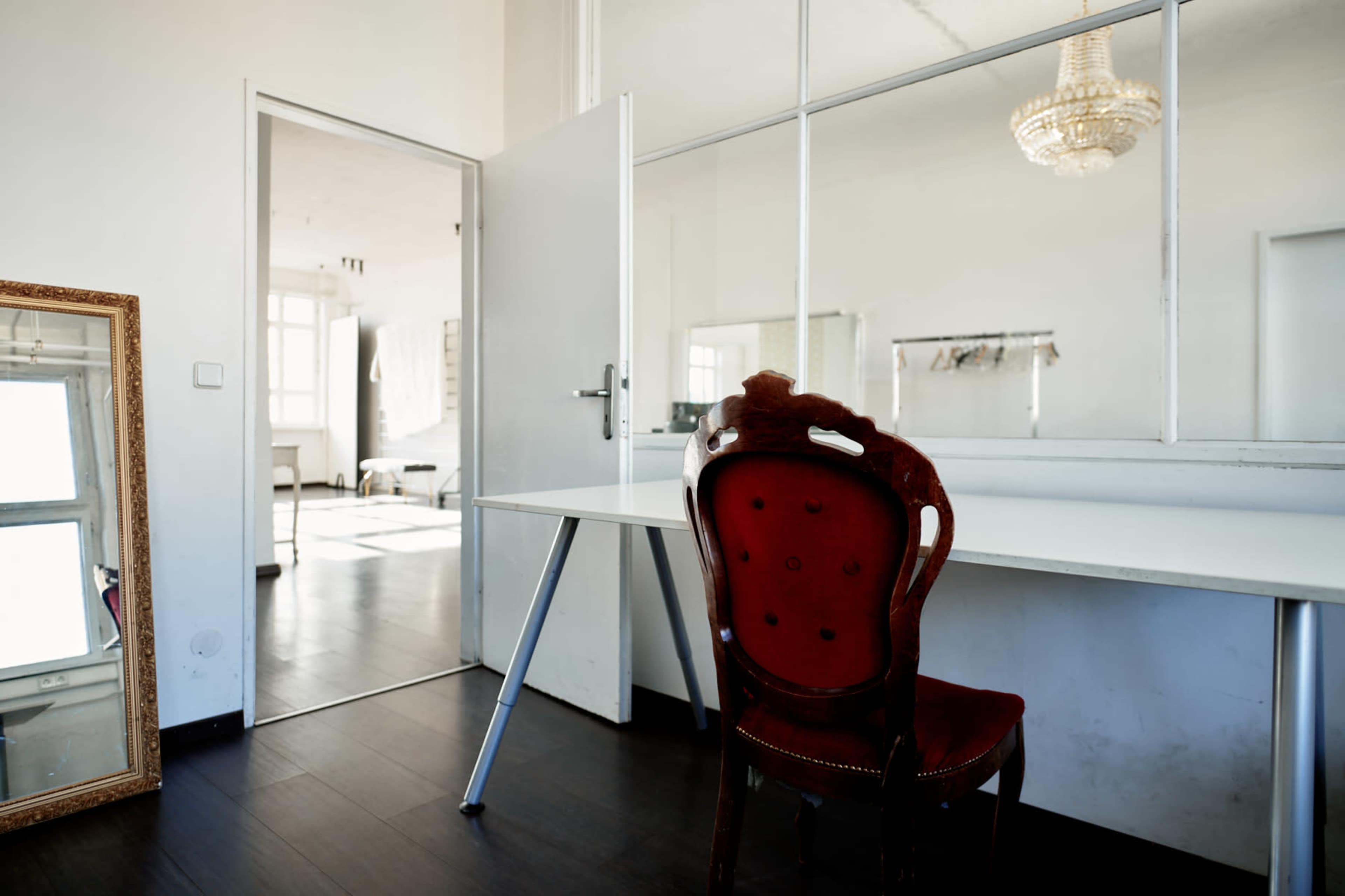 A room features a red upholstered chair at a white desk, with a mirror and a doorway leading to a well-lit open space in the background.