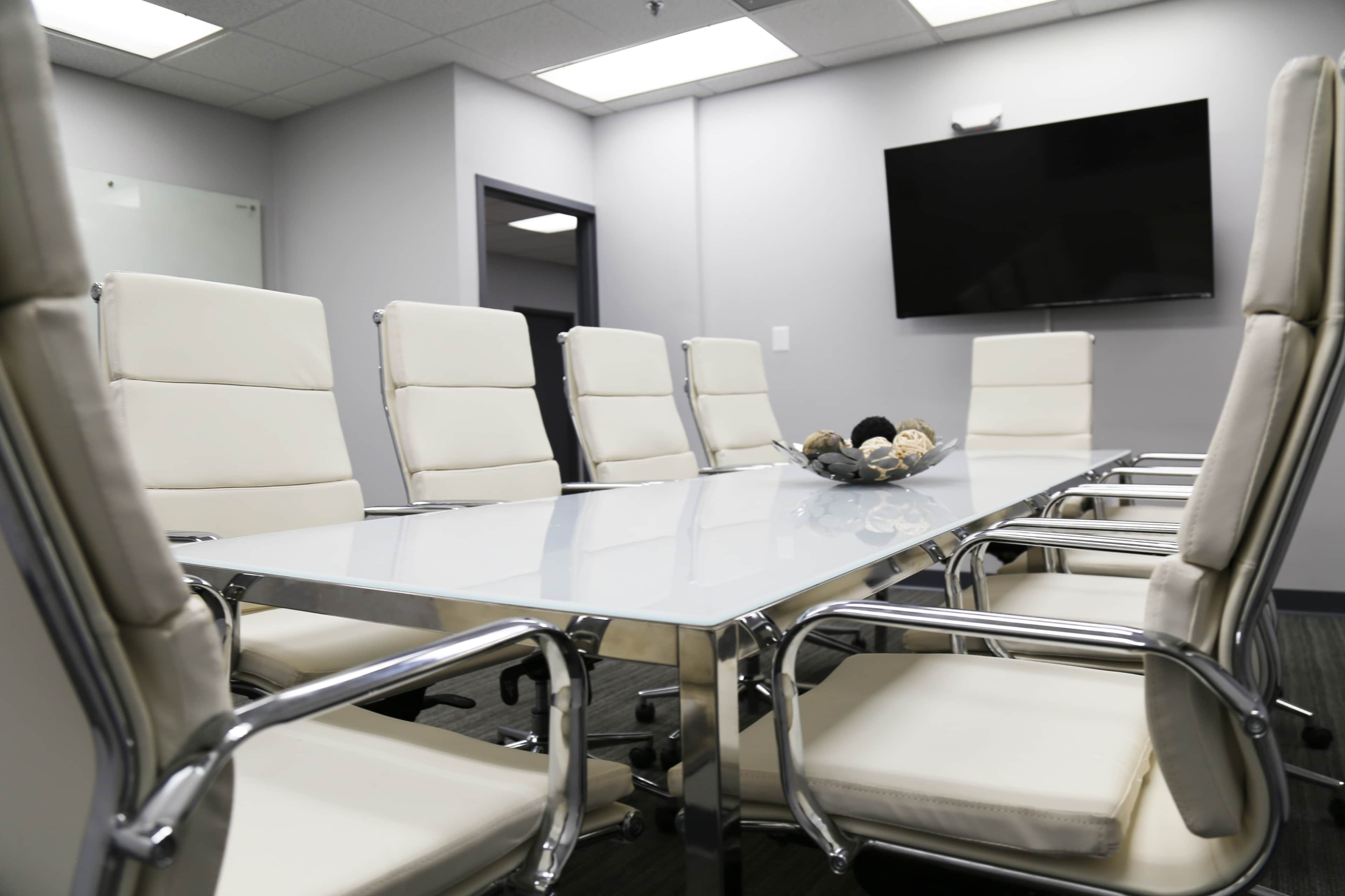 A conference room features a glass table surrounded by six white leather chairs and a wall-mounted television.