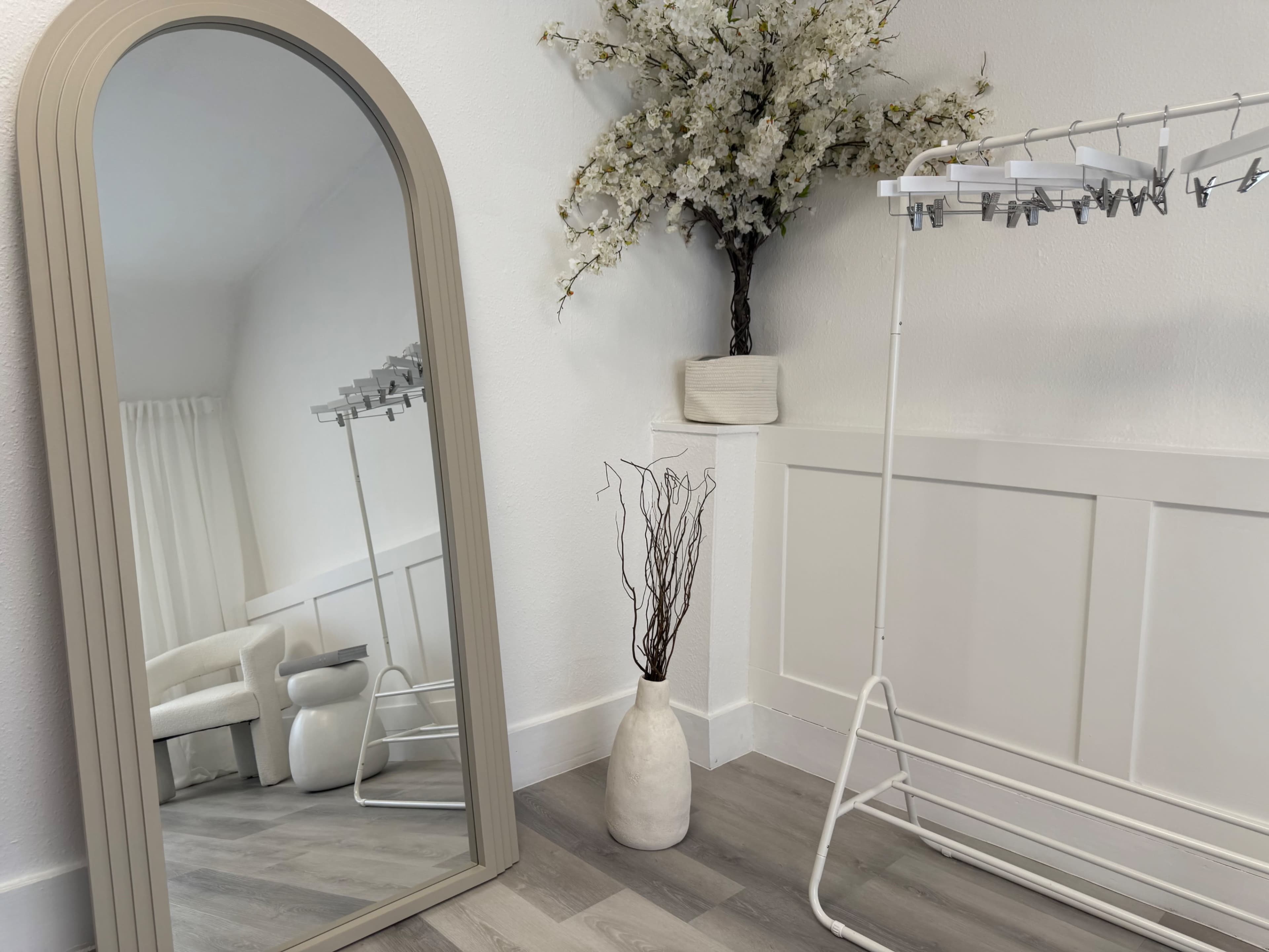 A large arched mirror reflects a minimalist room featuring a white chair, a vase with decorative branches, and a clothing rack against a white wall.