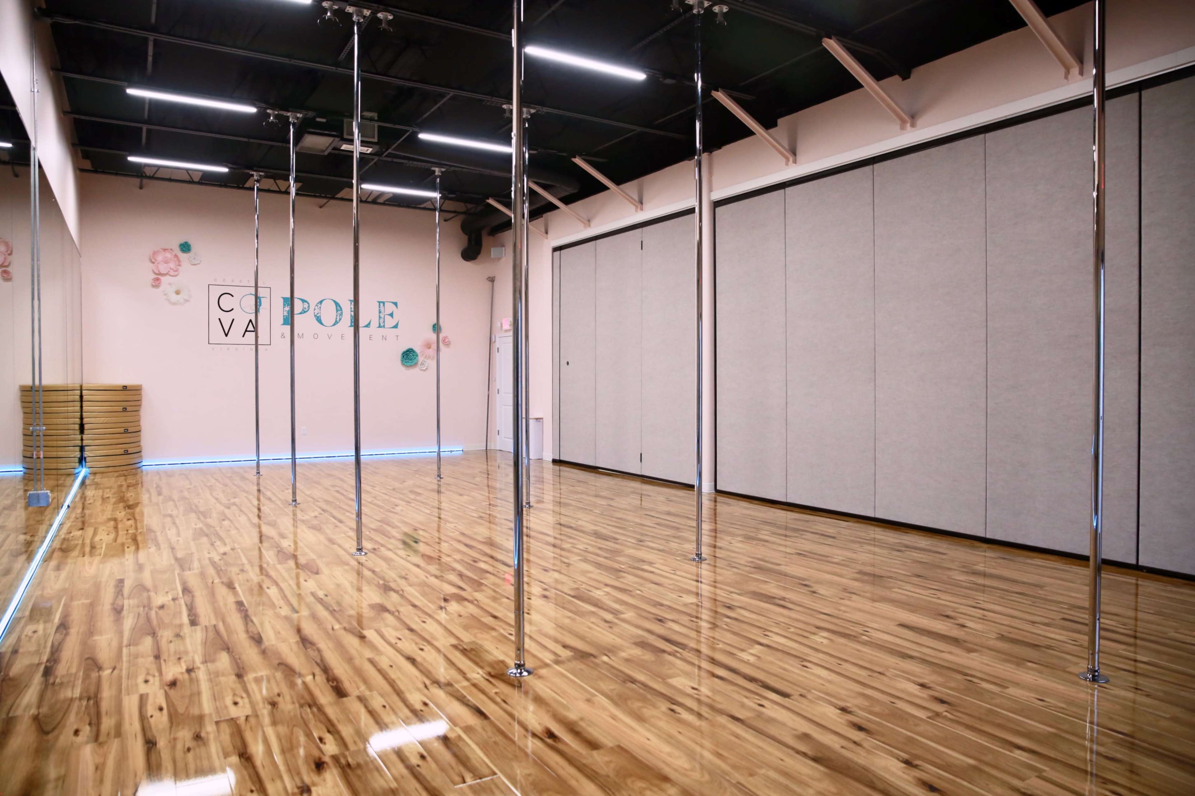 The image shows an indoor dance studio with shiny wooden flooring, several vertical poles, and a mirrored wall at the back.