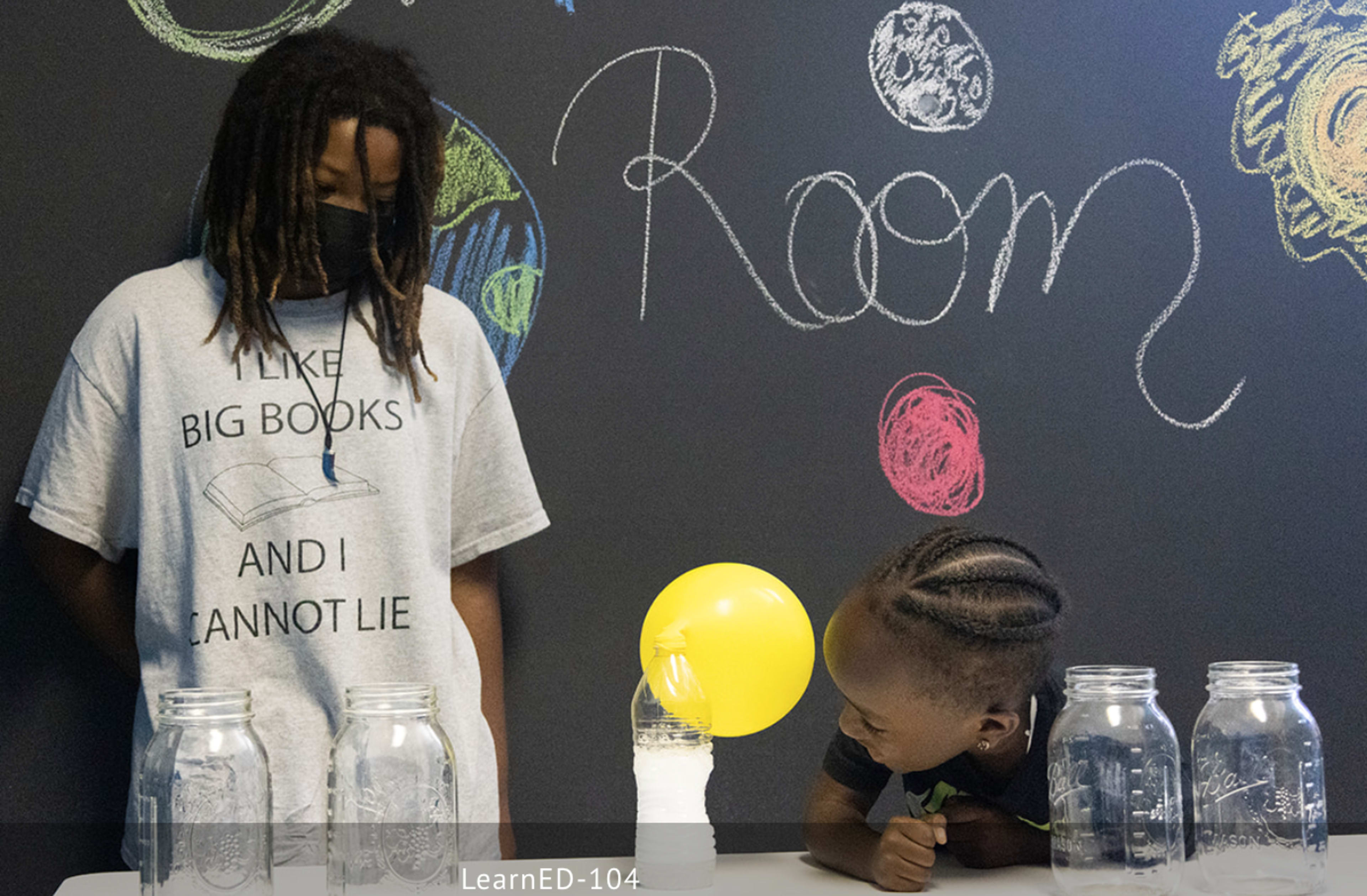 A child wearing a mask stands beside a table with glass jars while another child leans forward, looking at a balloon on the table in front of them, all set against a chalkboard wall with written text.