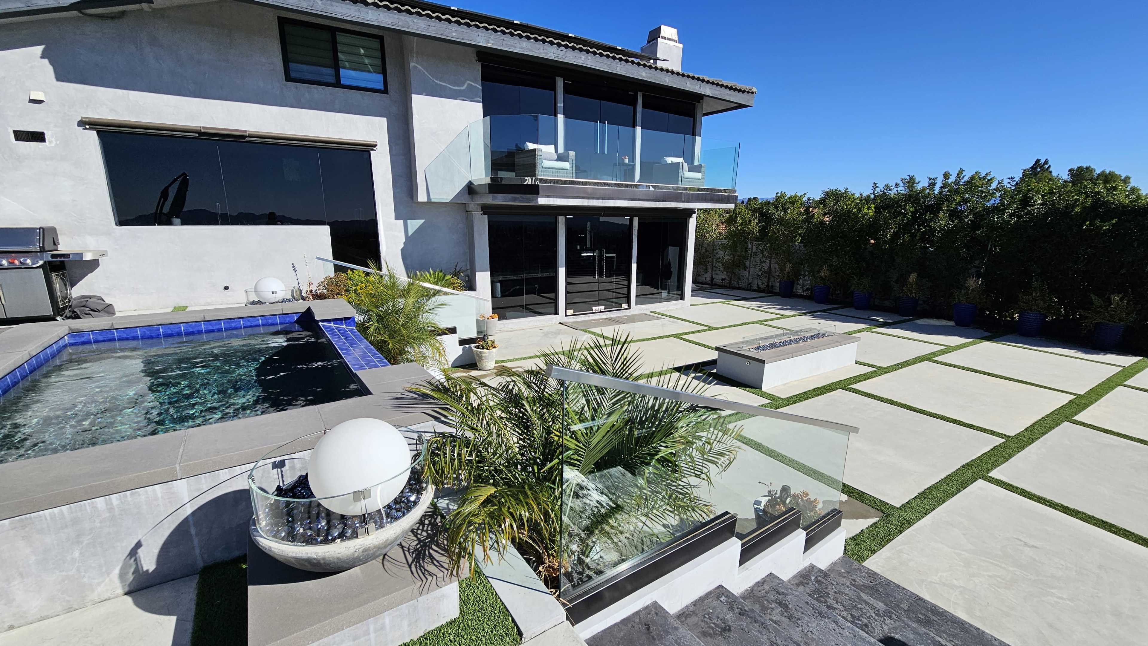 The image shows a modern outdoor space featuring a pool with a spa, landscaped areas, and a two-story house with large glass windows.
