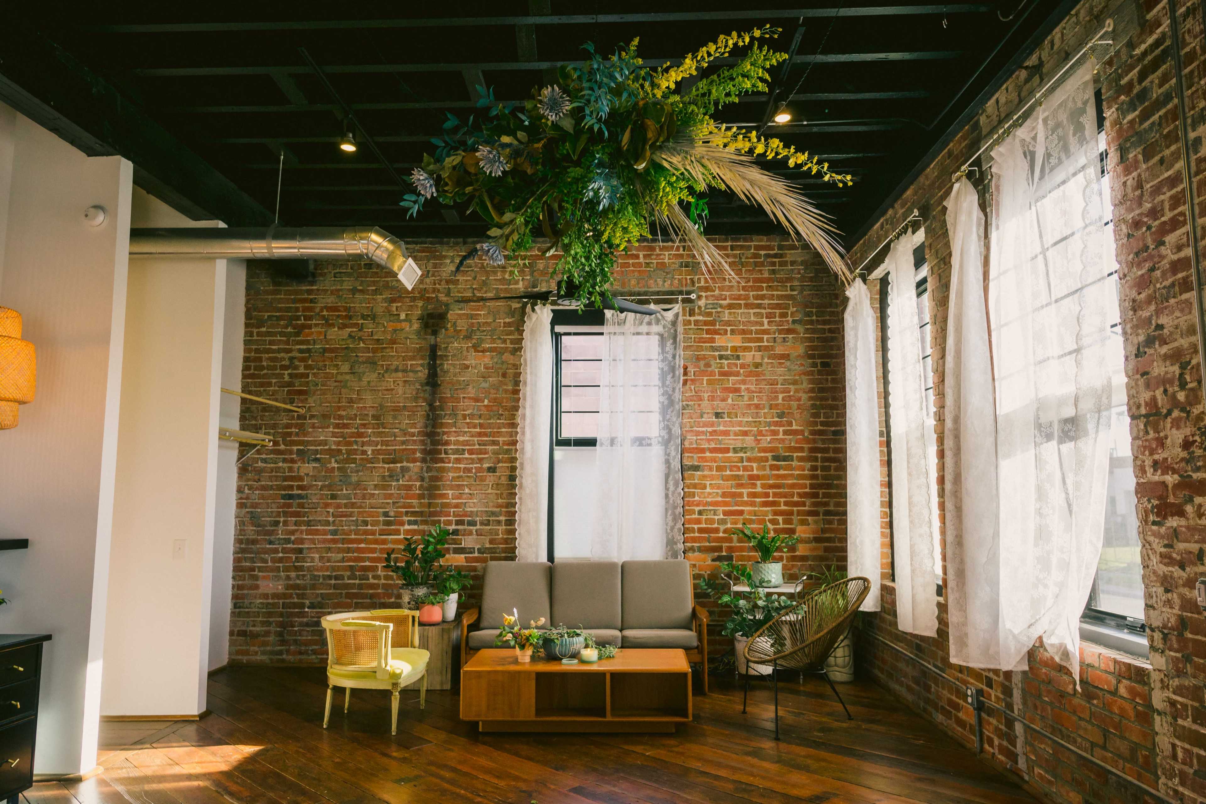 A spacious room with exposed brick walls, large windows adorned with sheer curtains, a sofa, a coffee table, and potted plants.