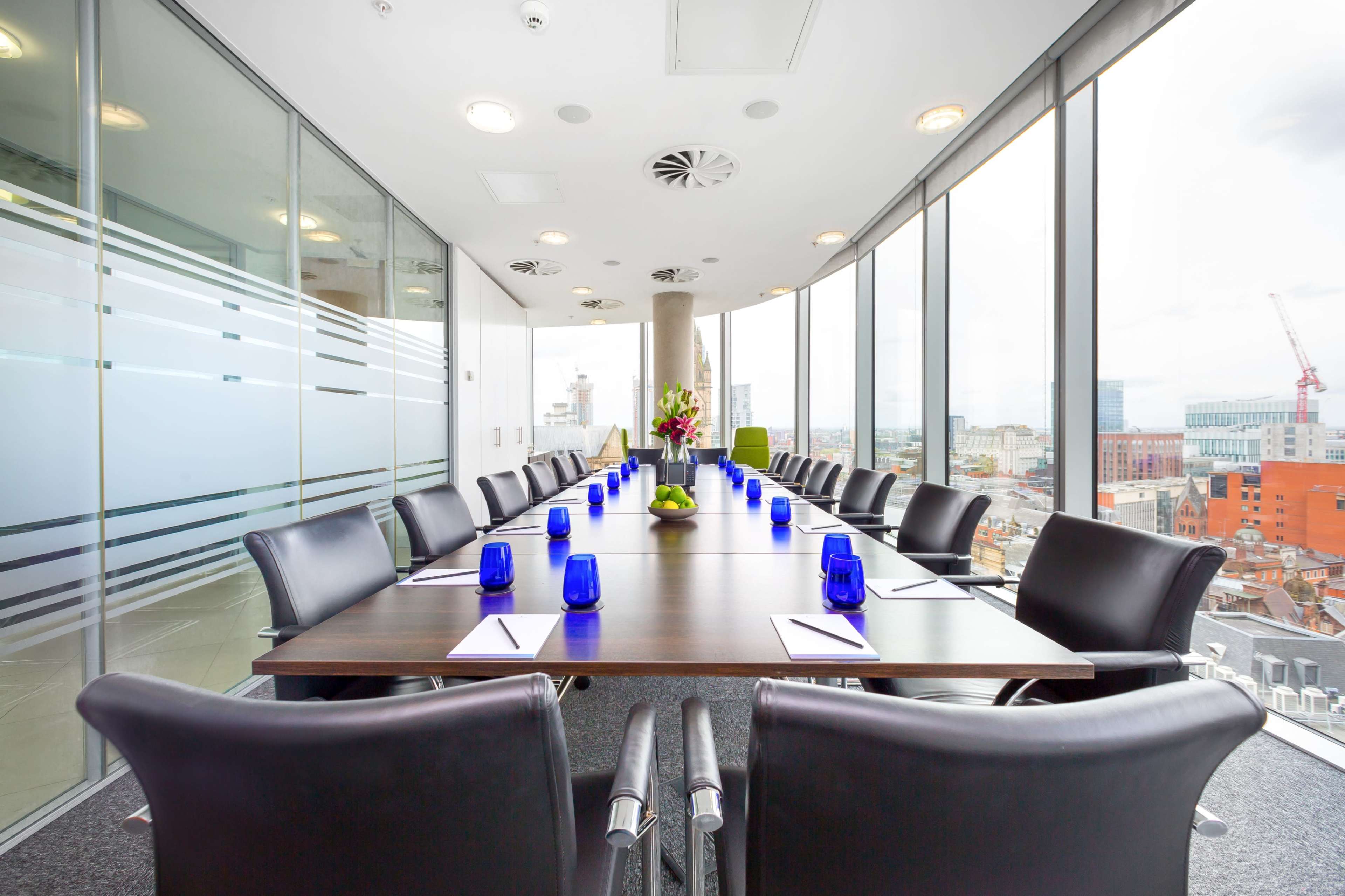 A boardroom with a large table surrounded by black chairs and blue centerpieces, featuring floor-to-ceiling windows that provide a view of a city skyline.