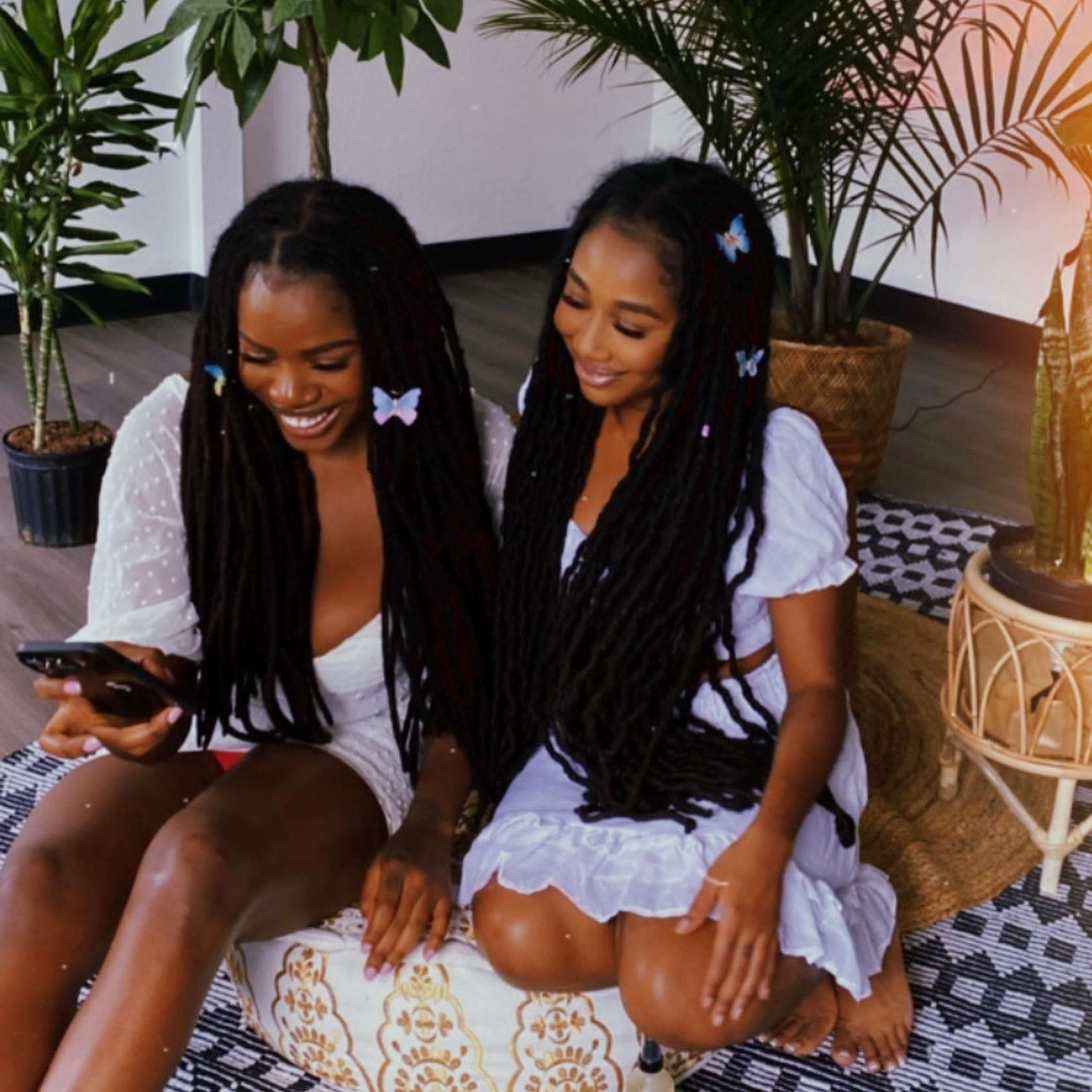 Two women with long, braided hair sit together on a woven rug, looking at a phone.