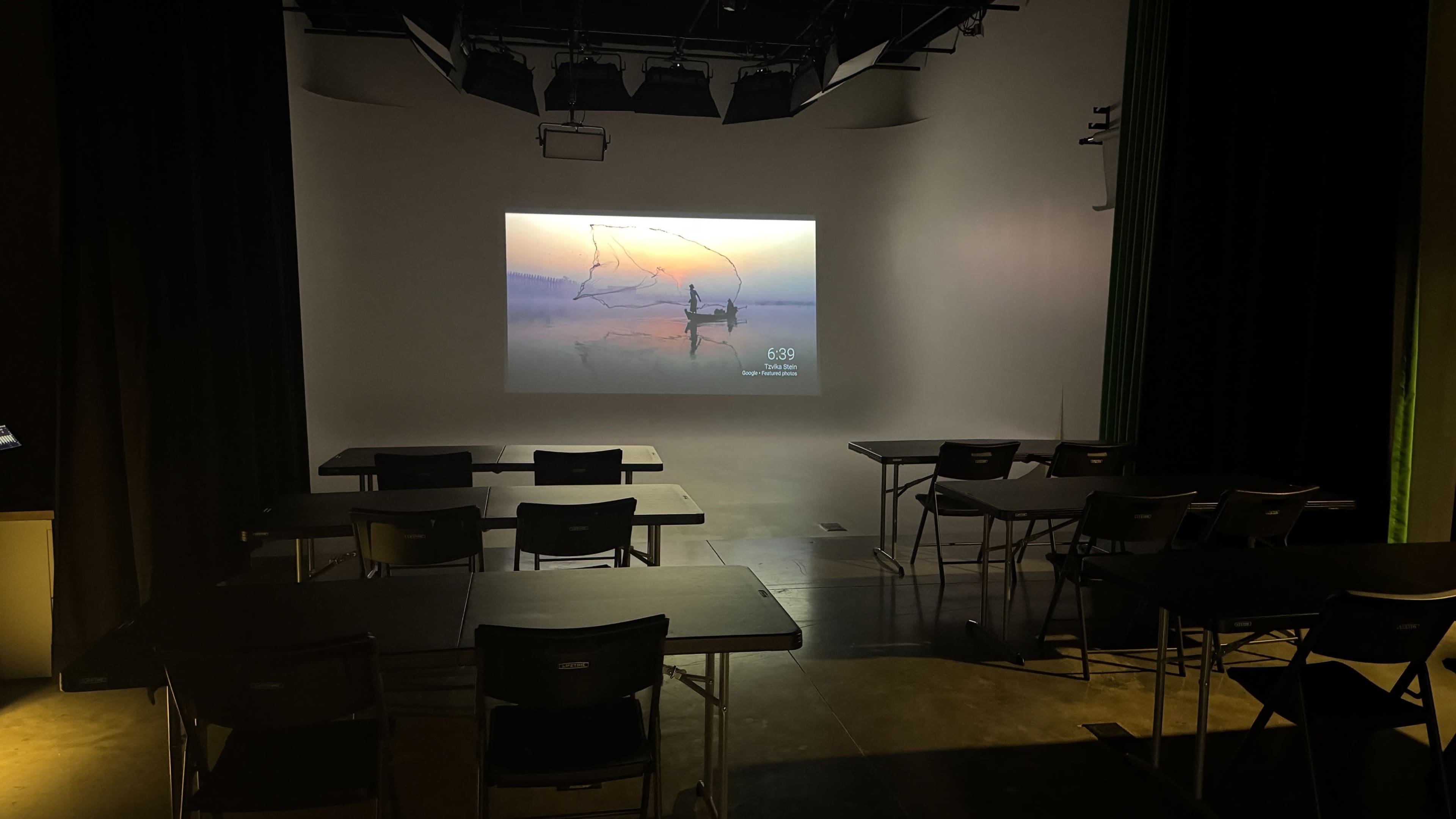 A blank projection screen displays an image of a person casting a net in a serene landscape, with several empty tables and chairs arranged in front of it in a dimly lit room.