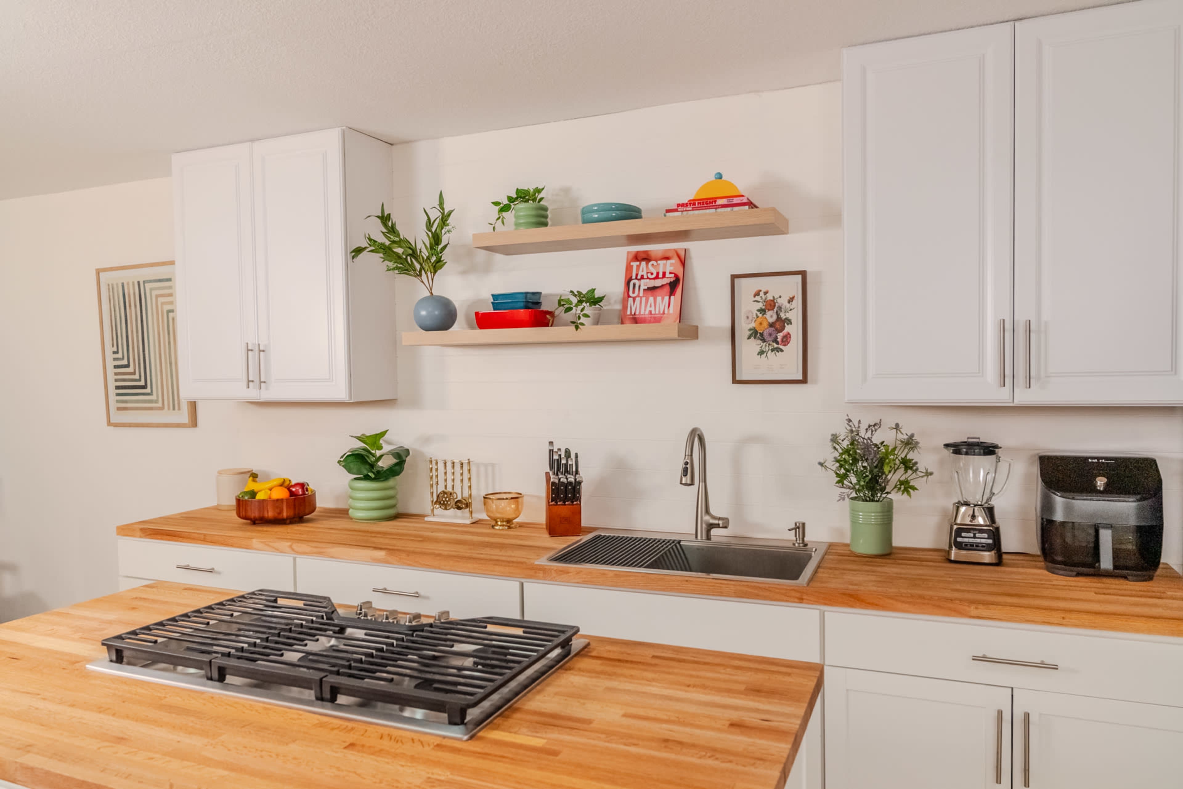 The image shows a modern kitchen with white cabinetry, a wooden countertop, and various decorative items and plants arranged on shelves.