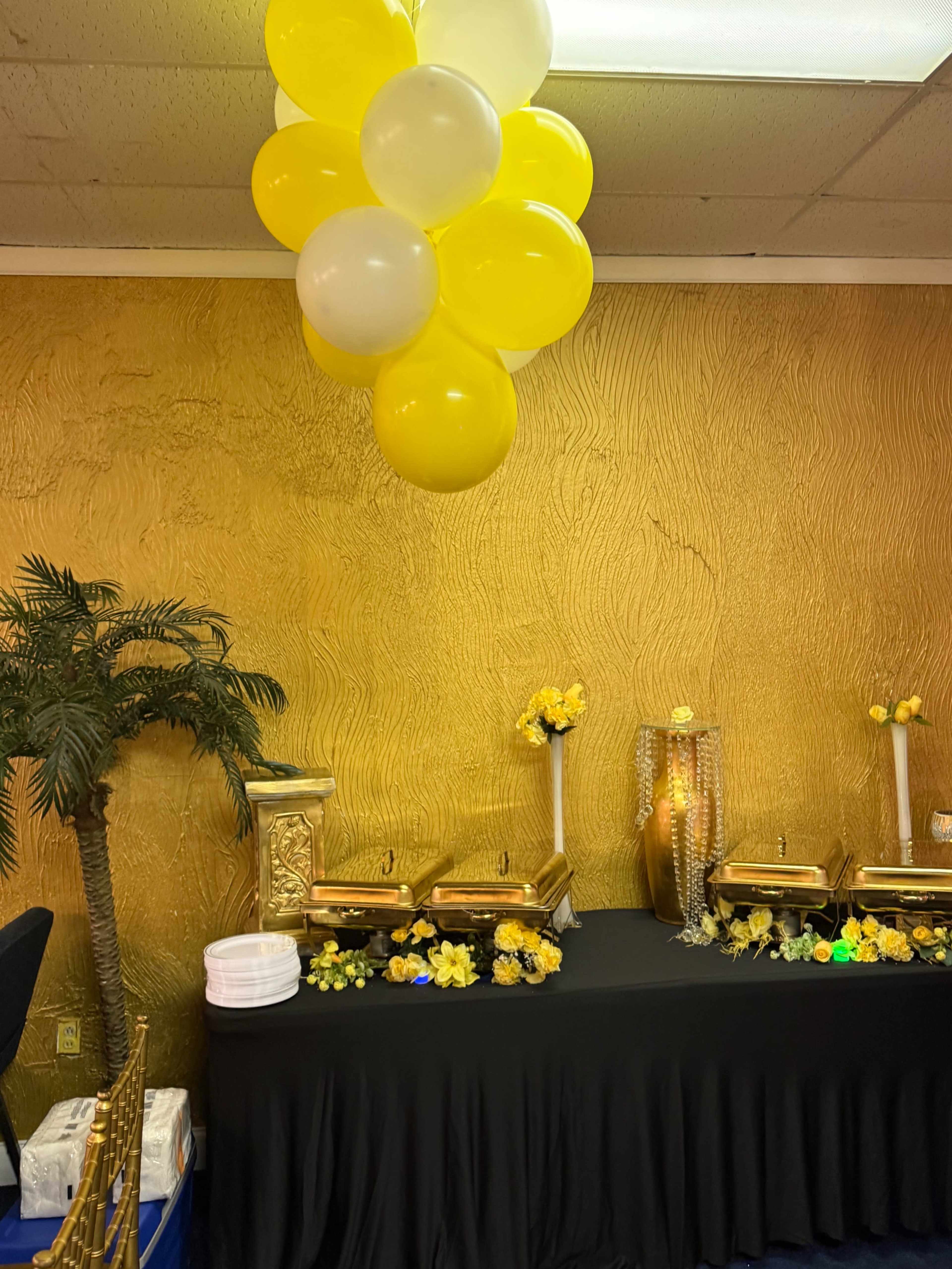 The image shows a buffet table decorated with black linens, yellow flowers, and gold chafing dishes, beneath a cluster of yellow and white balloons against a textured golden wall.