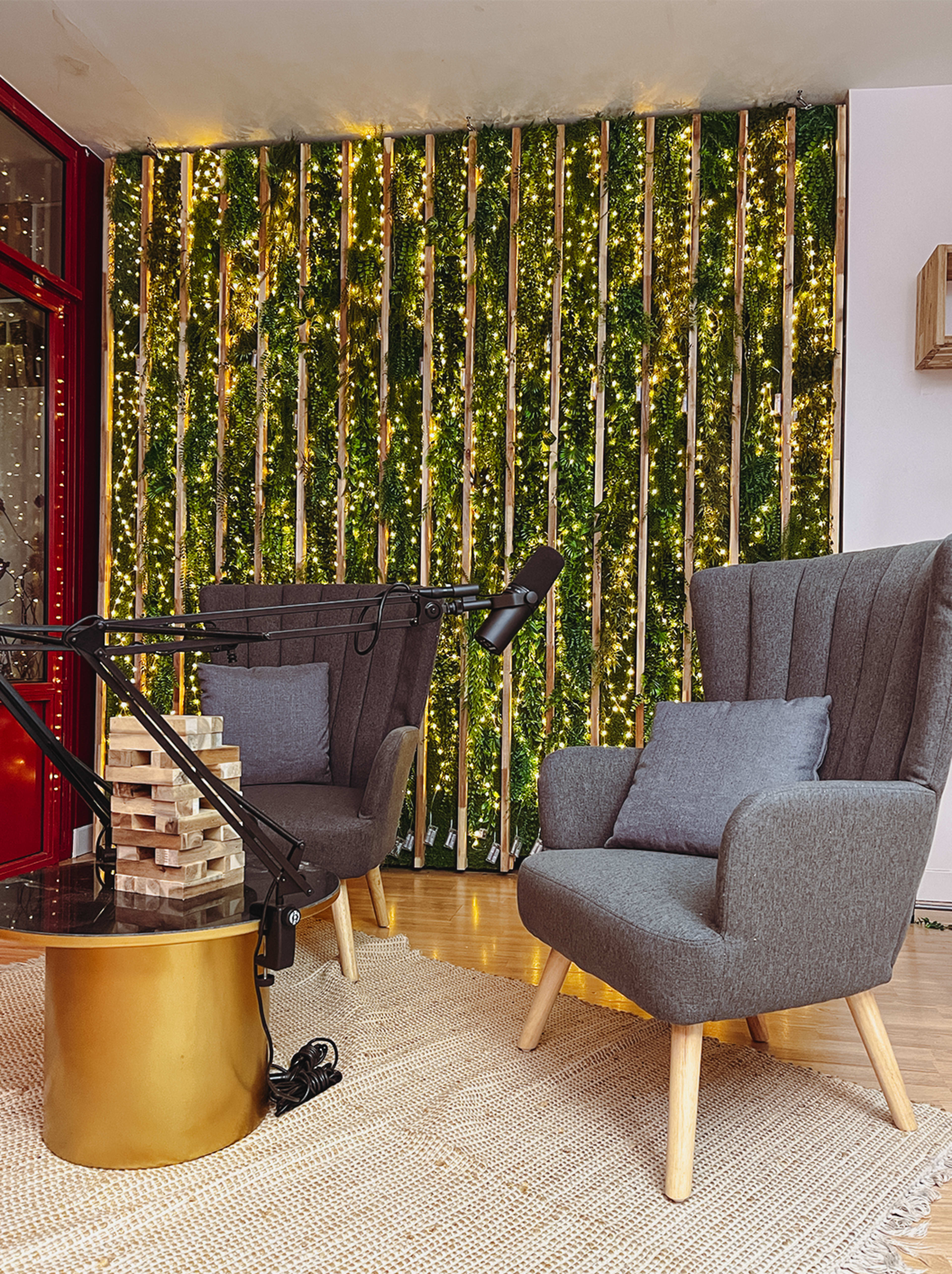 A cozy seating area features two gray armchairs beside a small round gold table, with a microphone on an adjustable arm and a backdrop of greenery adorned with string lights.
