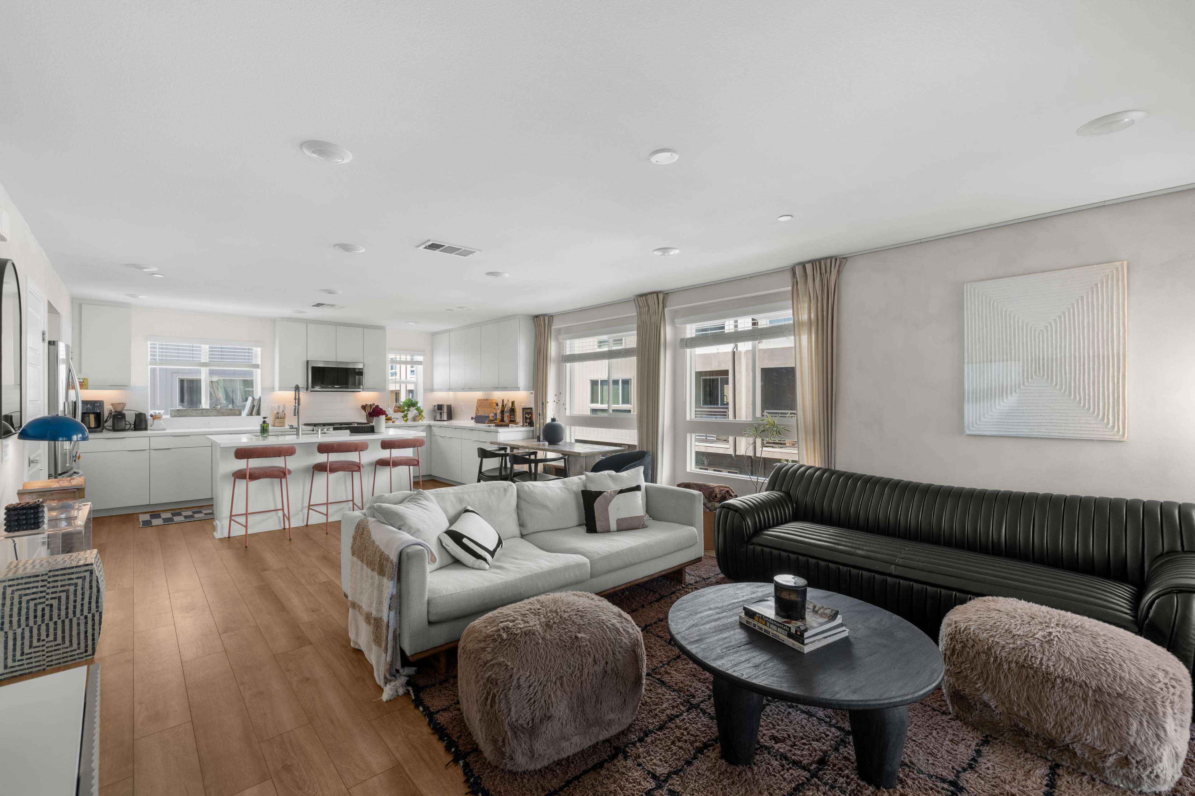 The image features a modern living space with a kitchen and living area, showcasing a sofa, armchairs, a round coffee table, and large windows that allow natural light to enter.