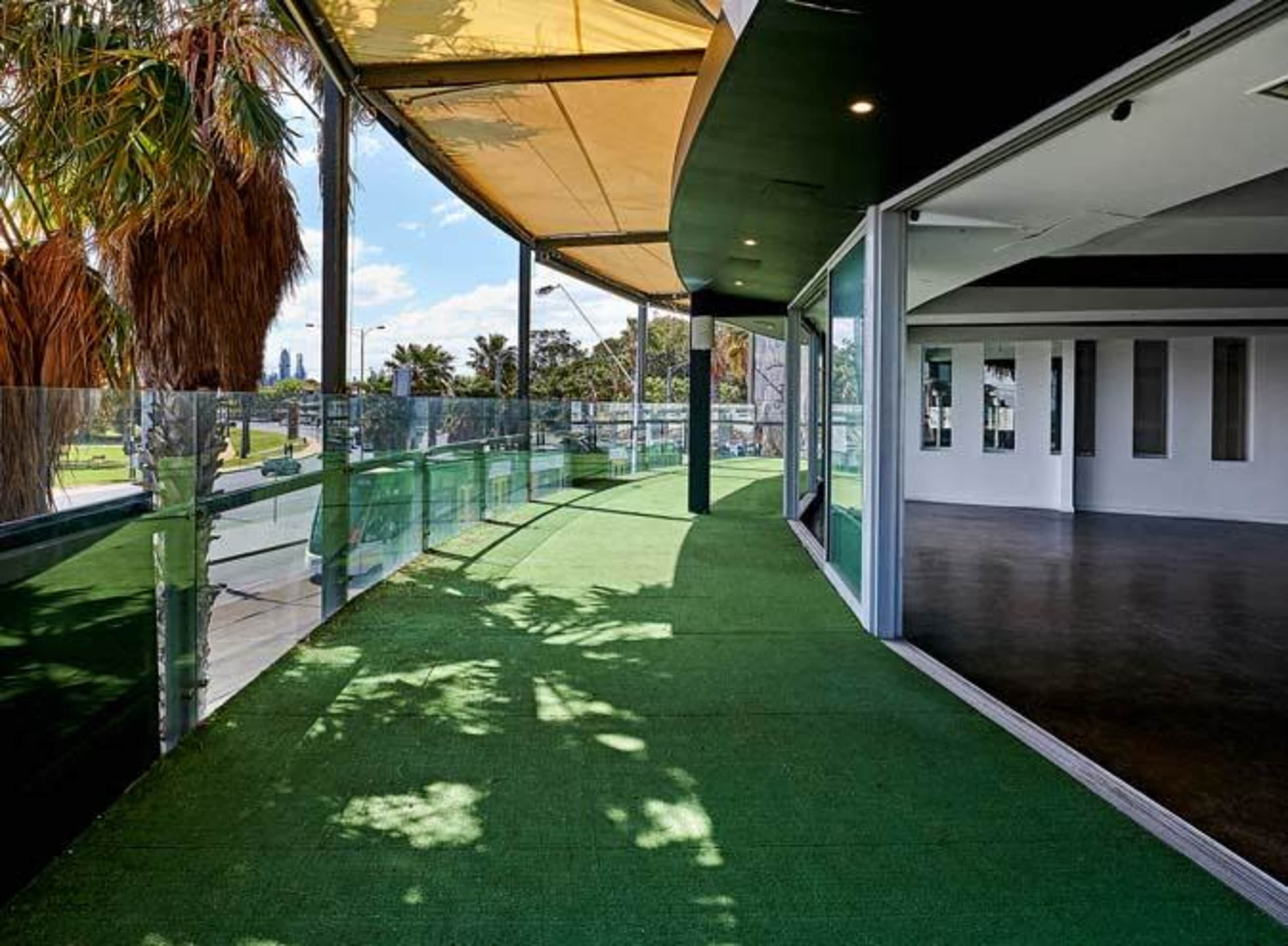 A modern balcony area with green turf flooring, surrounded by glass railings and a partially shaded roof.