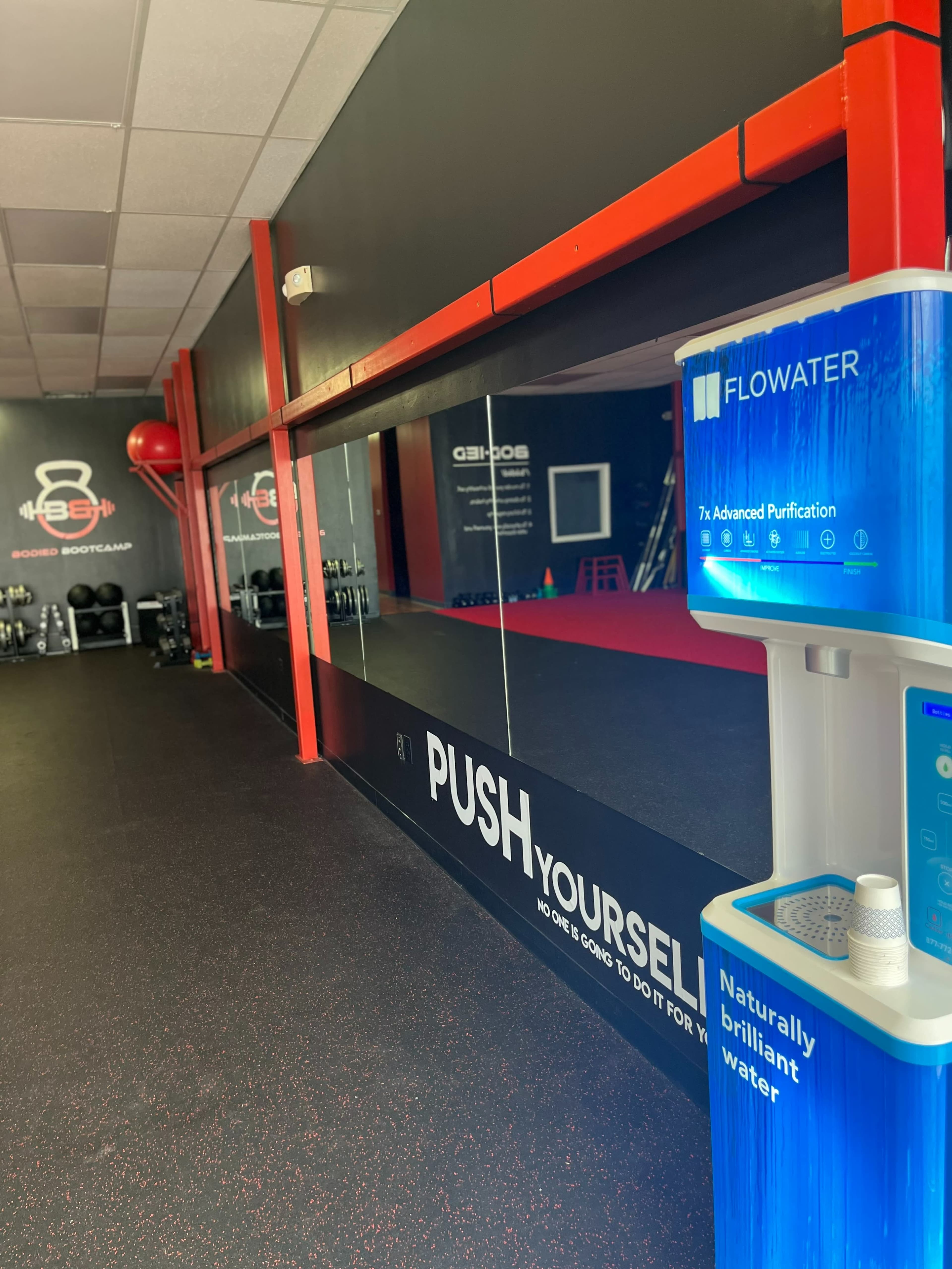 The image shows the interior of a gym with a large mirror, a water refill station, and motivational text on the wall.