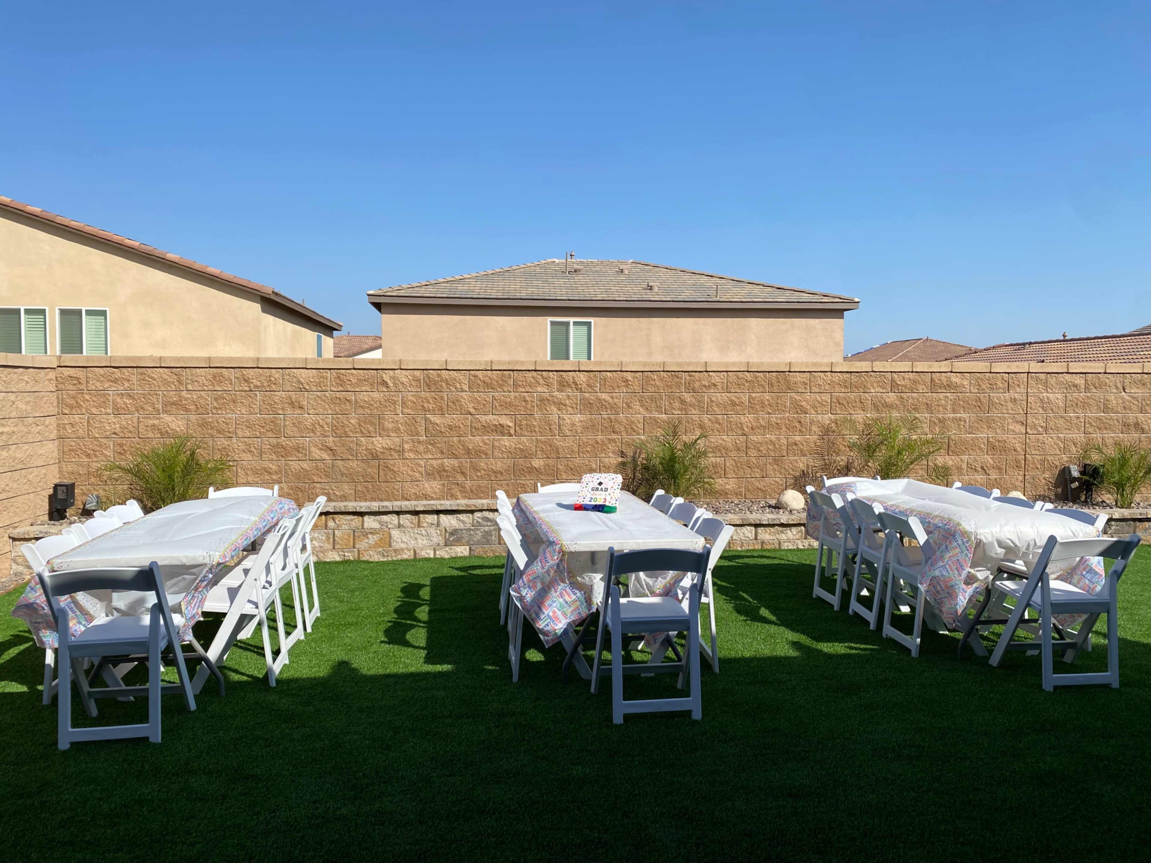 A backyard space is set up with multiple tables covered in white tablecloths, arranged neatly on green grass, and flanked by beige walls of nearby houses.