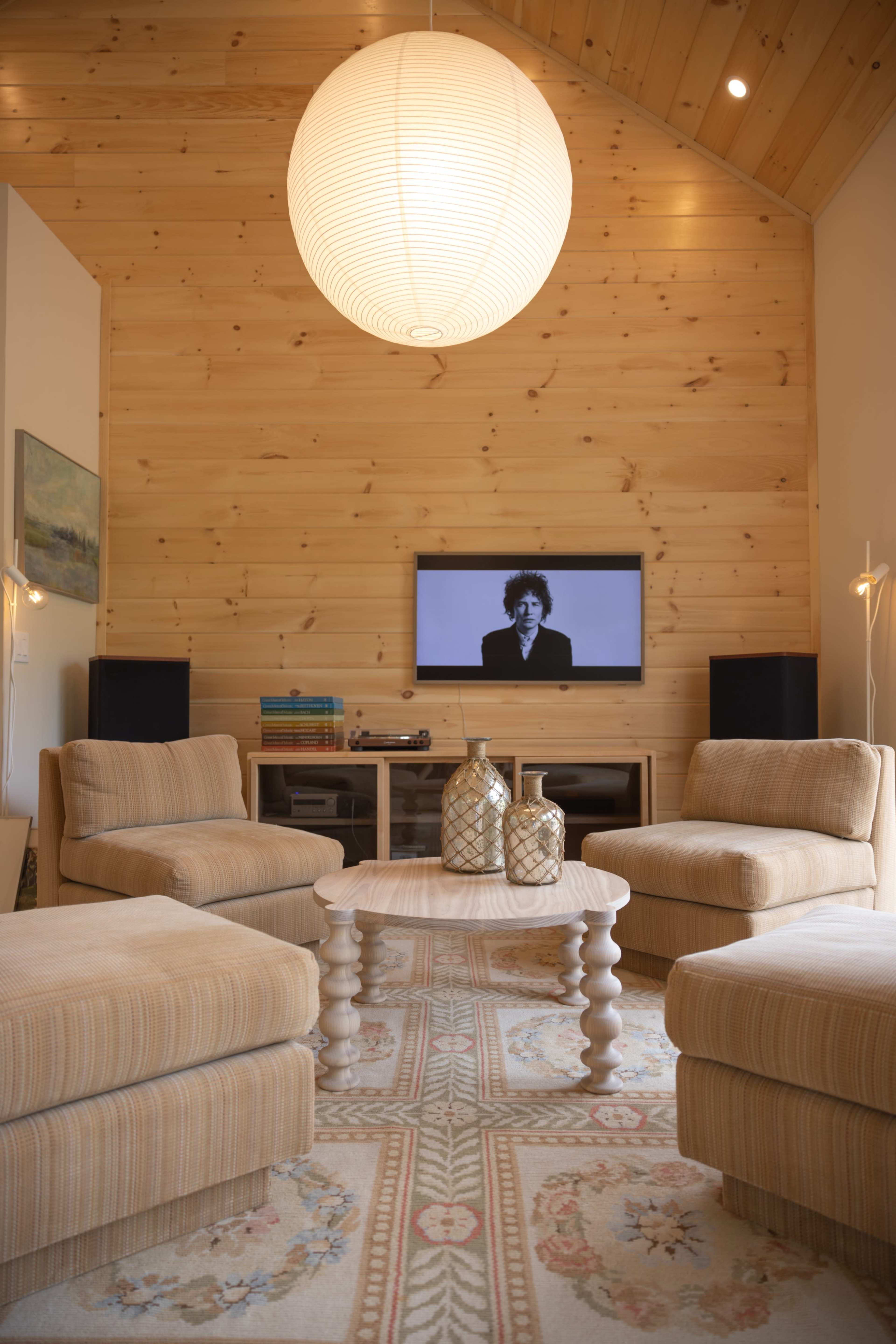 A modern living room features two striped armchairs, a round wooden coffee table, and a television mounted on a wooden wall, illuminated by a large paper lantern overhead.