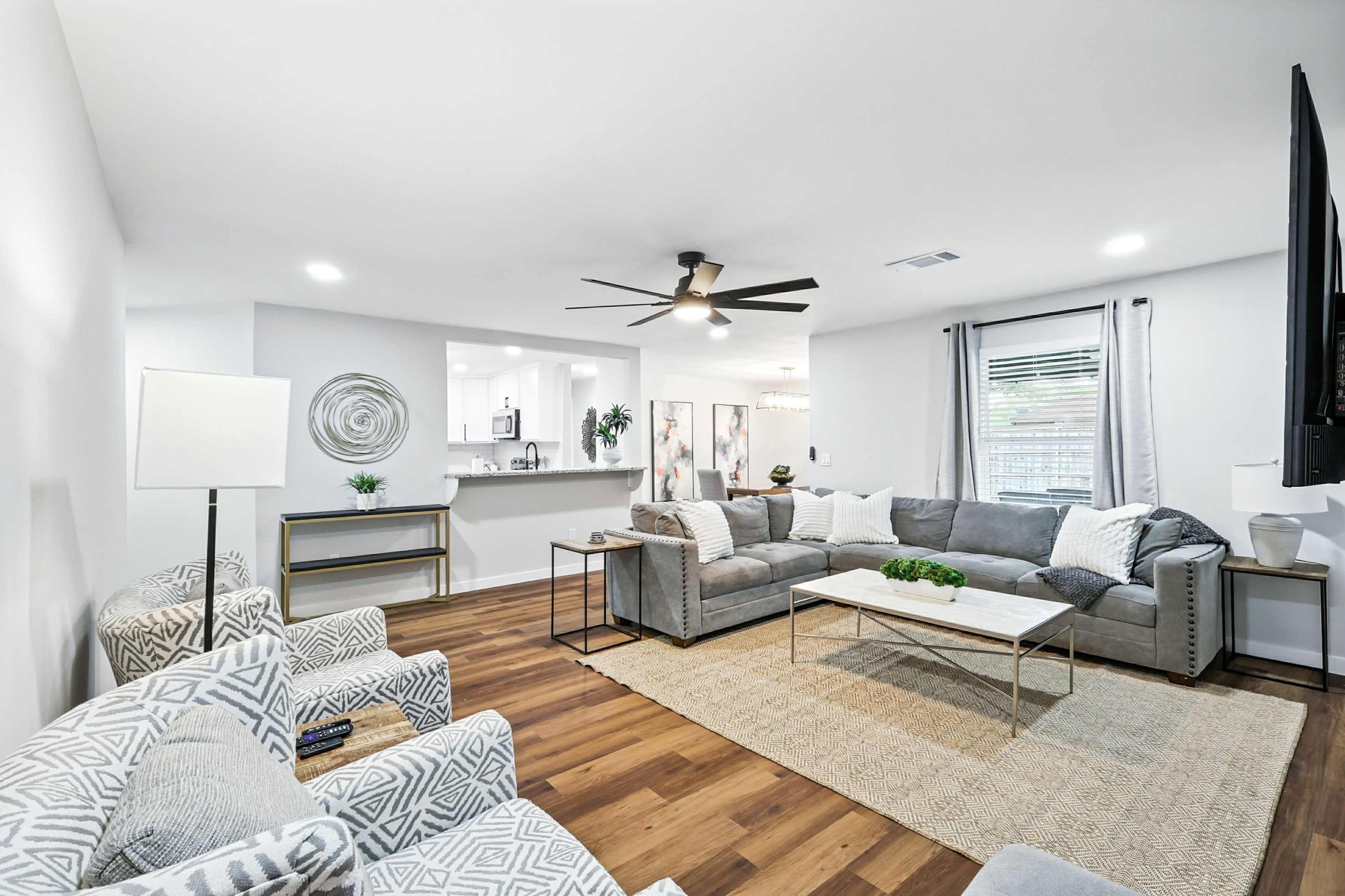 The living room features a gray sectional sofa, a coffee table, and two patterned armchairs, with a ceiling fan and a light-colored area rug on hardwood flooring.