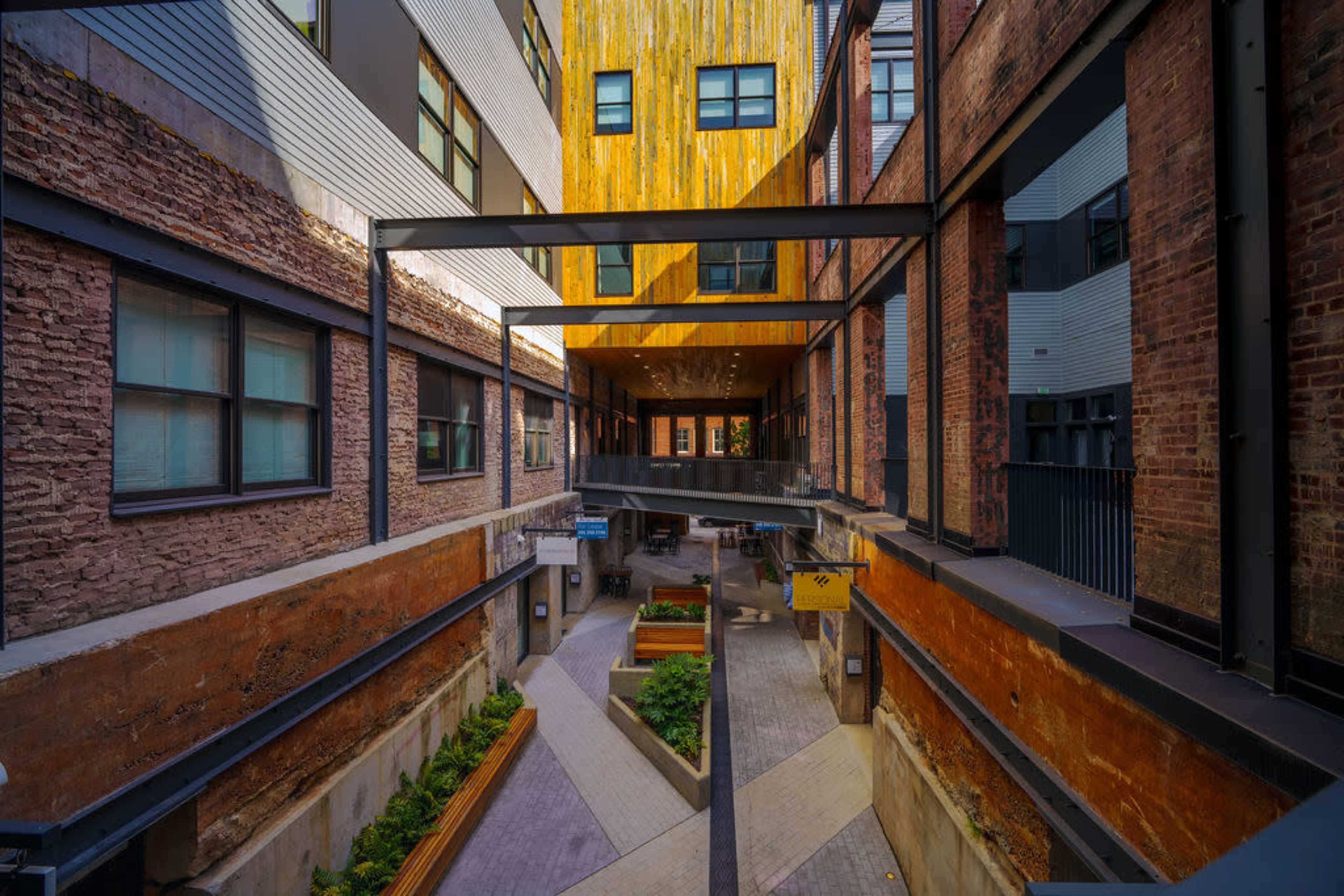 The image shows a modern urban courtyard featuring brick walls, wooden accents, and a pathway lined with greenery.