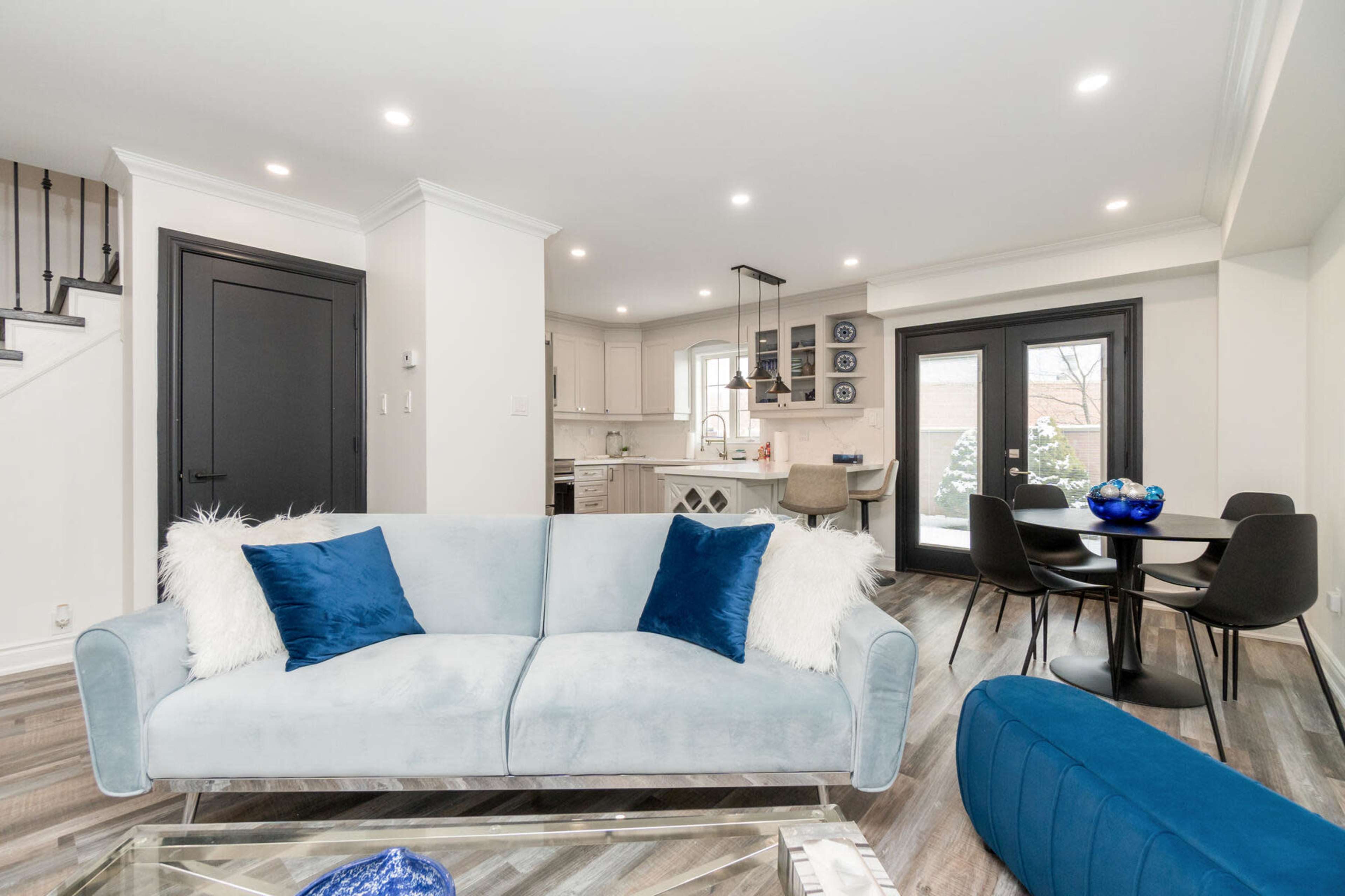 A modern living space features a light blue sofa with decorative pillows, a dining area with black chairs, and a kitchen visible in the background.
