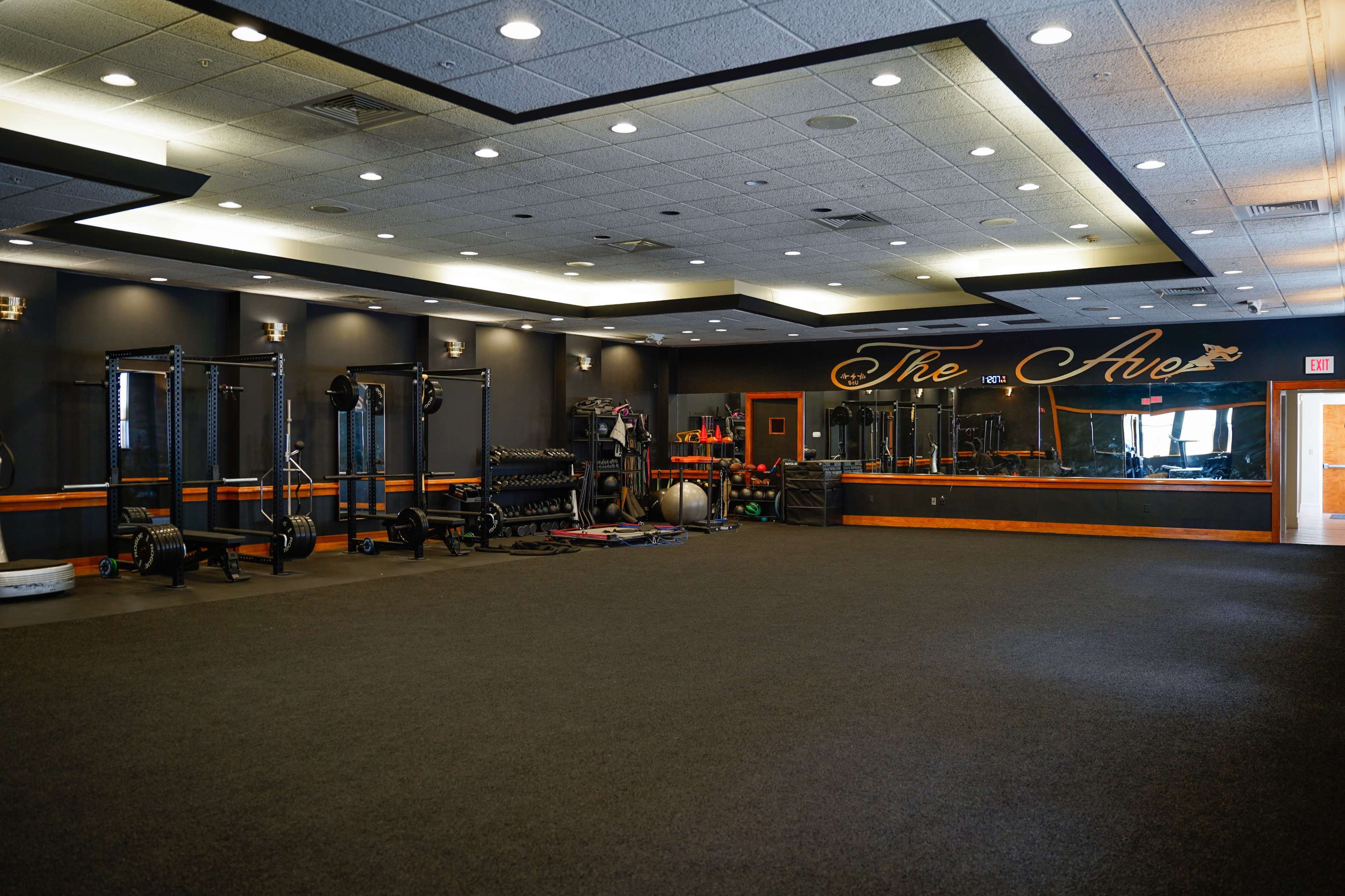 An empty gym interior featuring black walls, workout equipment, and a mirrored wall with "The Ave" written on it.