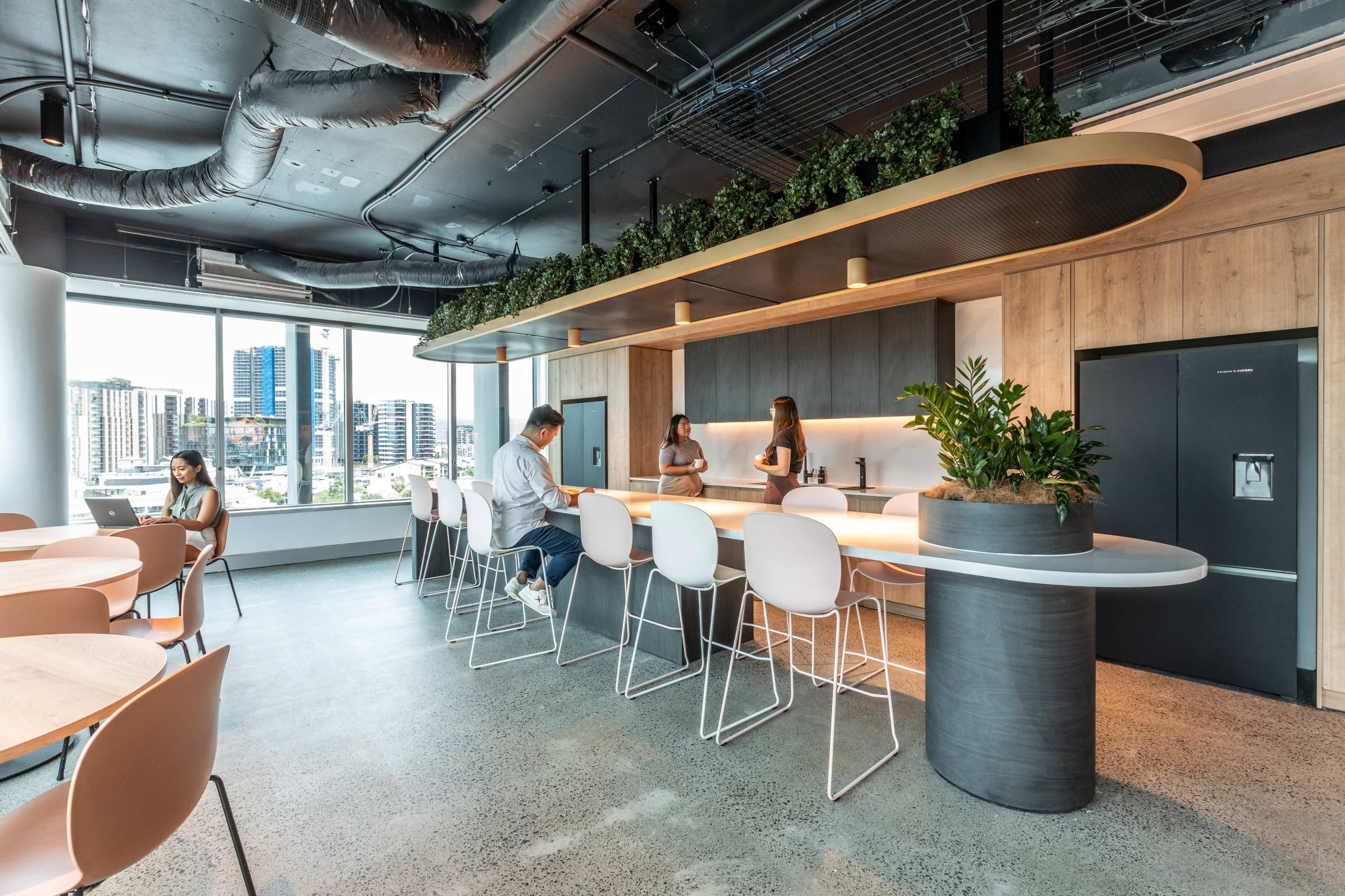 A modern office break room features a large island counter with seating, plants along the ceiling, and large windows overlooking a cityscape.