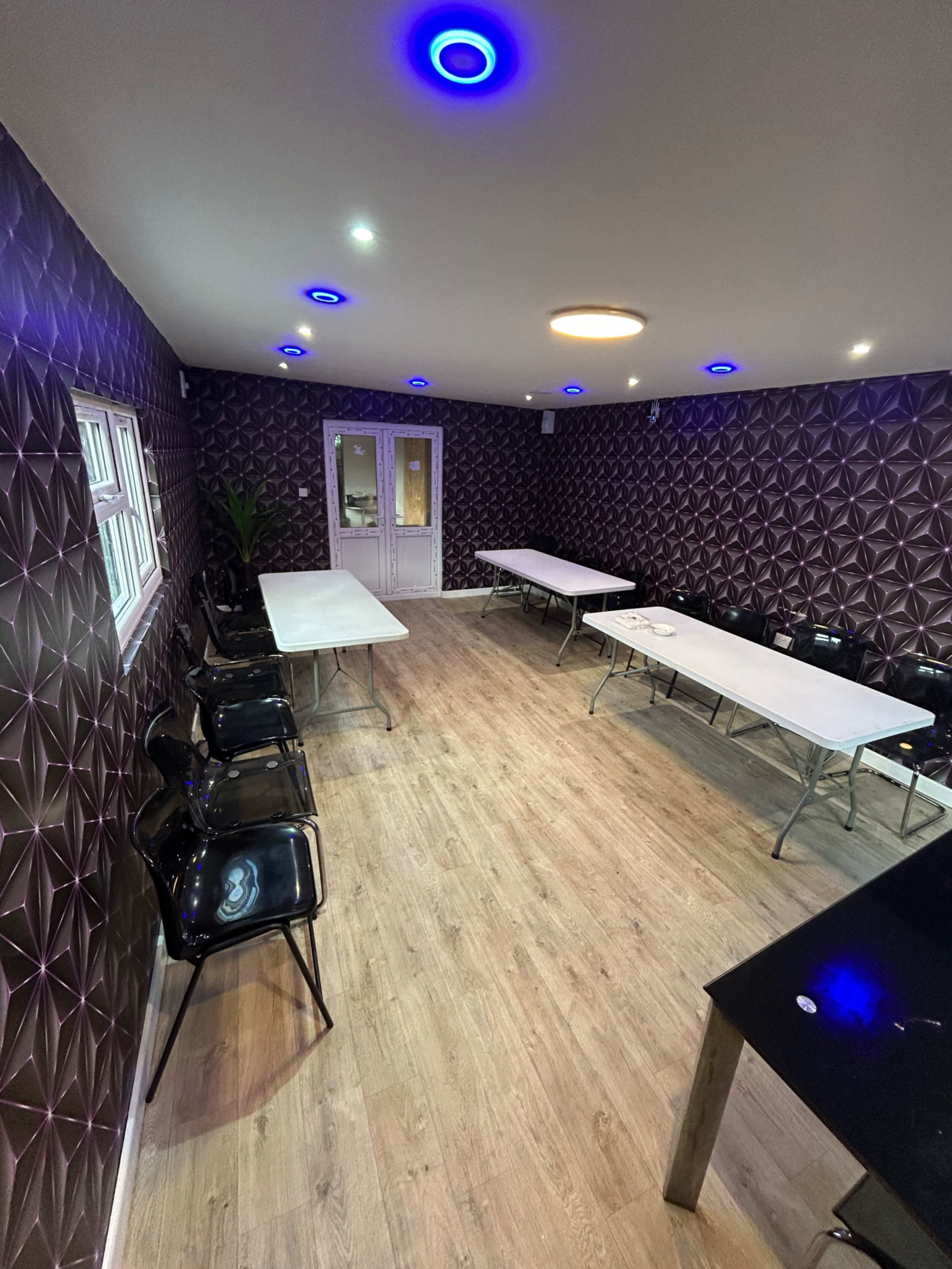 The image shows a spacious room with patterned walls, several long tables arranged, and black chairs positioned around them under blue and white ceiling lights.