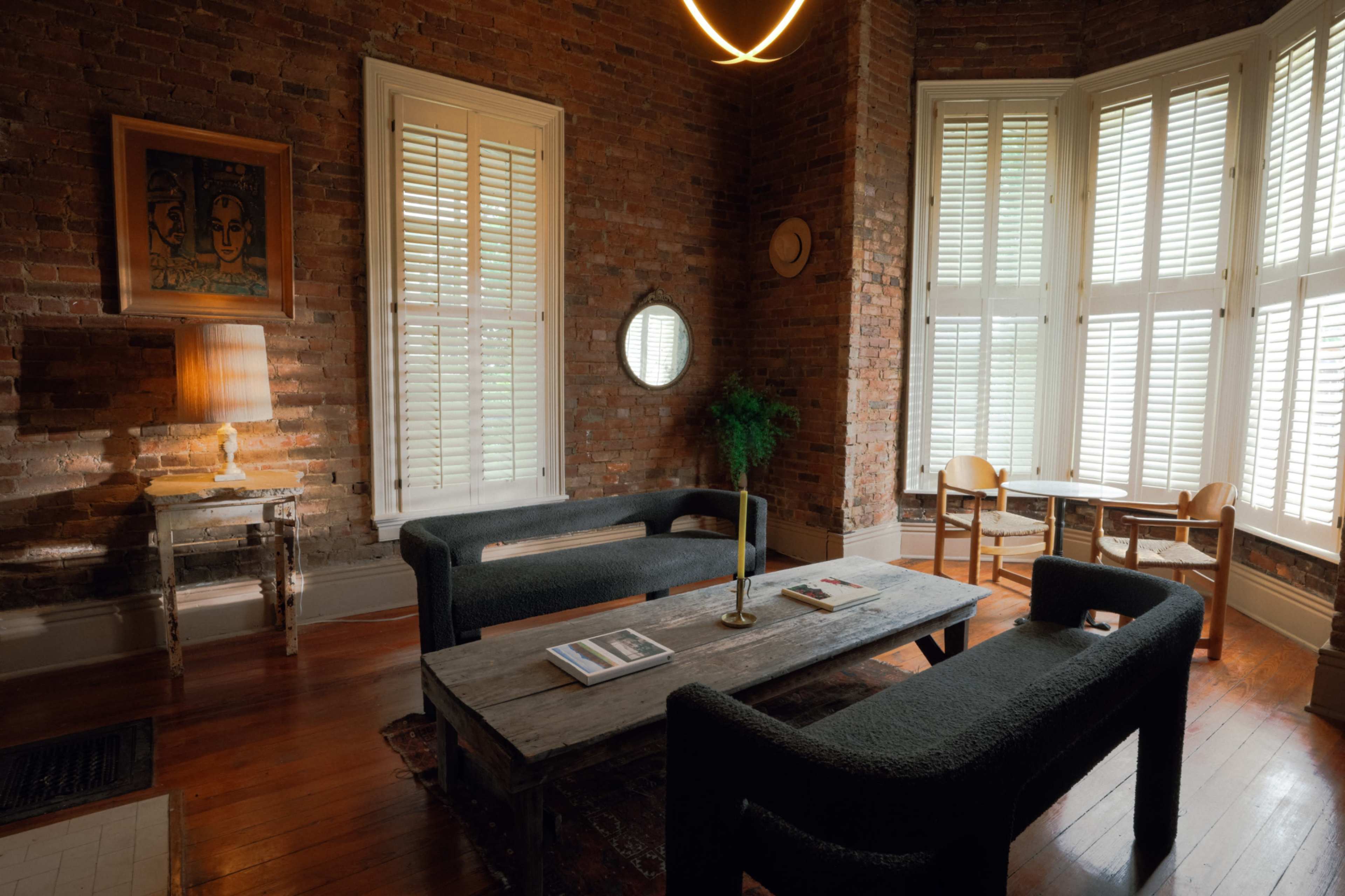 The room features exposed brick walls, wooden flooring, and minimalistic furniture, including two dark sofas, a wooden coffee table, and a small dining area with chairs.