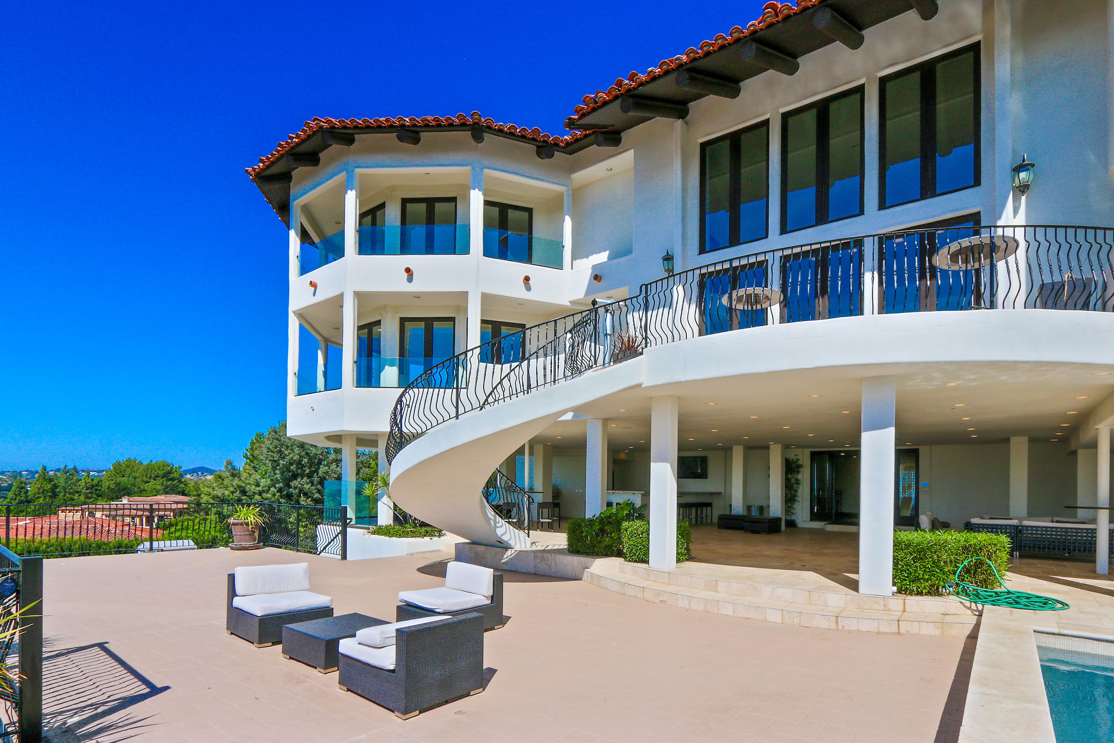 A modern, multi-level house with large windows and a curved balcony, overlooking a patio area with outdoor seating.