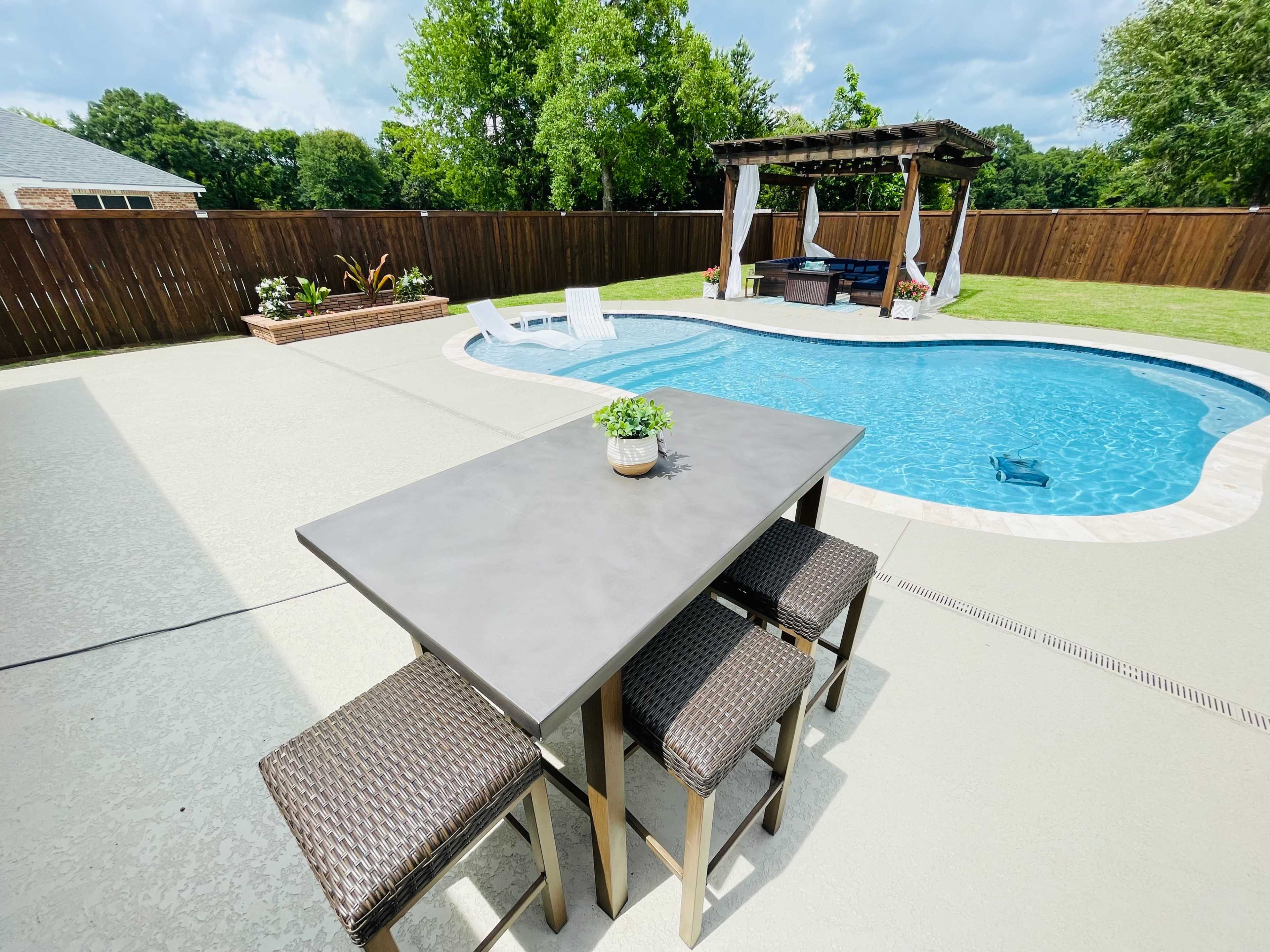 A concrete table with four chairs sits beside a swimming pool surrounded by a wooden fence and a pergola.