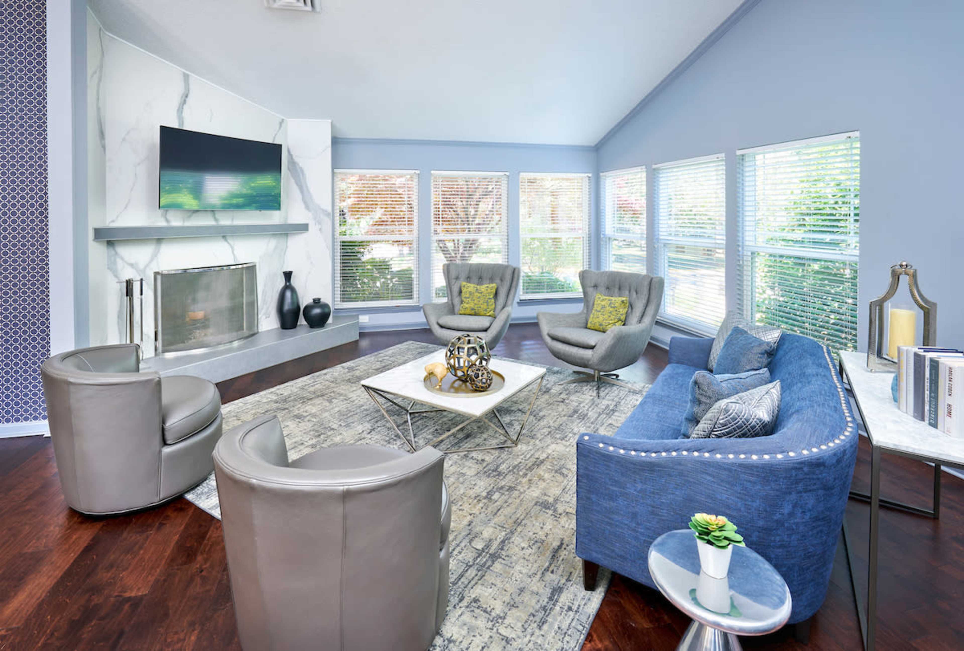 A modern living room features two gray chairs and a blue couch arranged around a coffee table, with large windows providing natural light and a marble fireplace as a focal point.