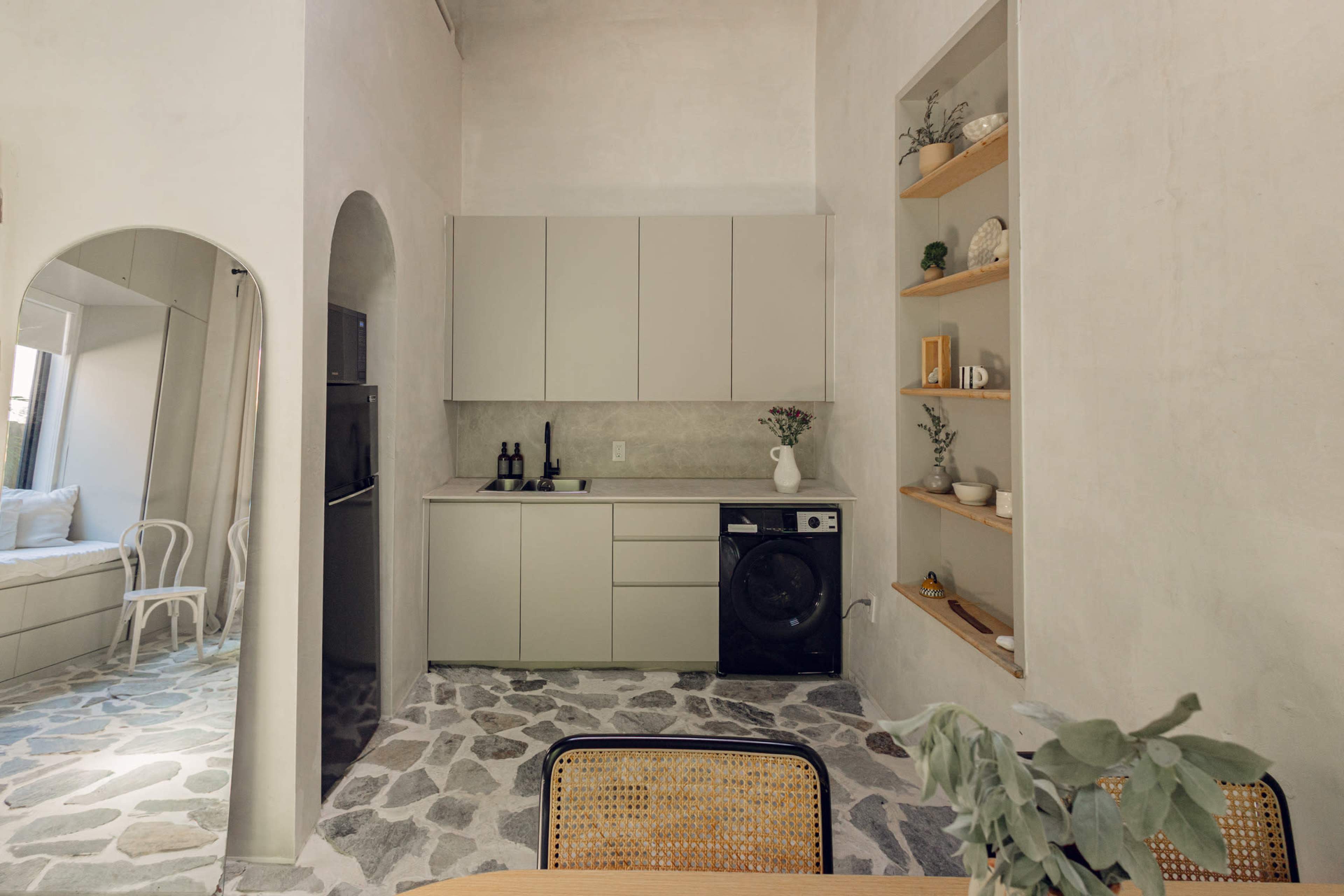 The image shows a minimalist kitchen with light-colored cabinets, a dark washing machine, and open shelves containing various items, all set in a space with stone flooring.