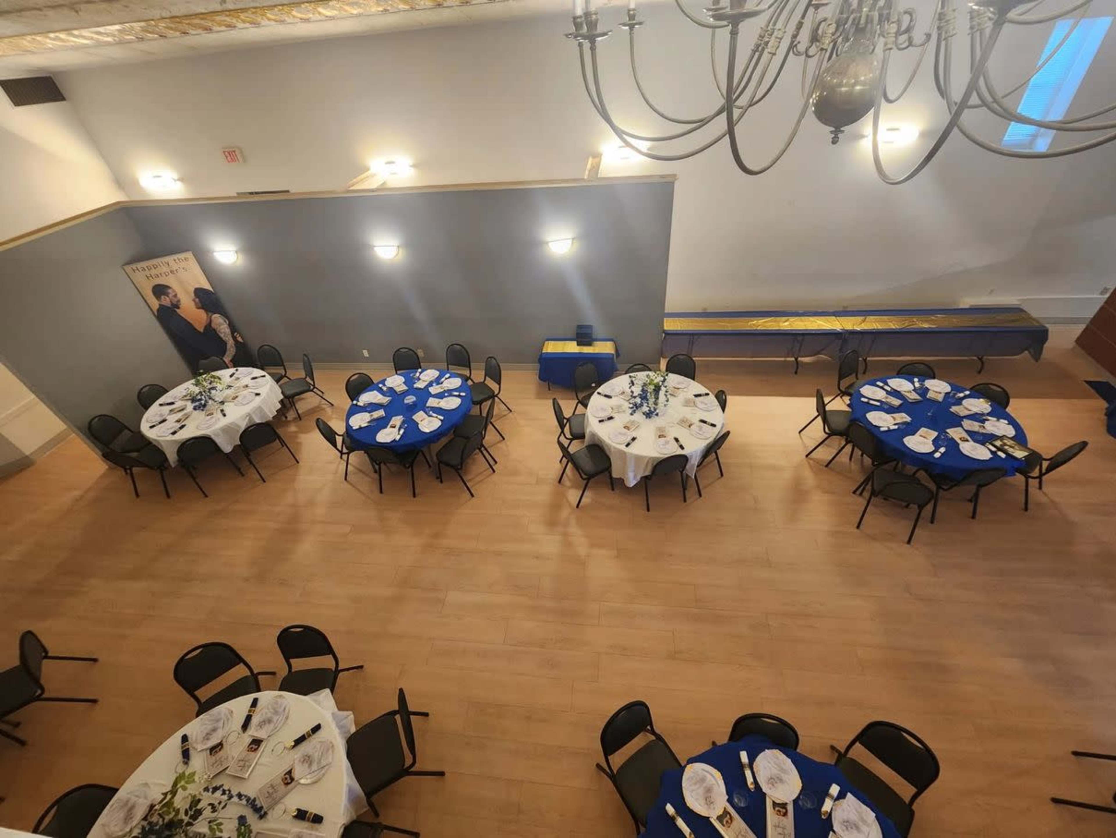 A spacious event hall features round tables with blue tablecloths set for a meal, illuminated by chandeliers.