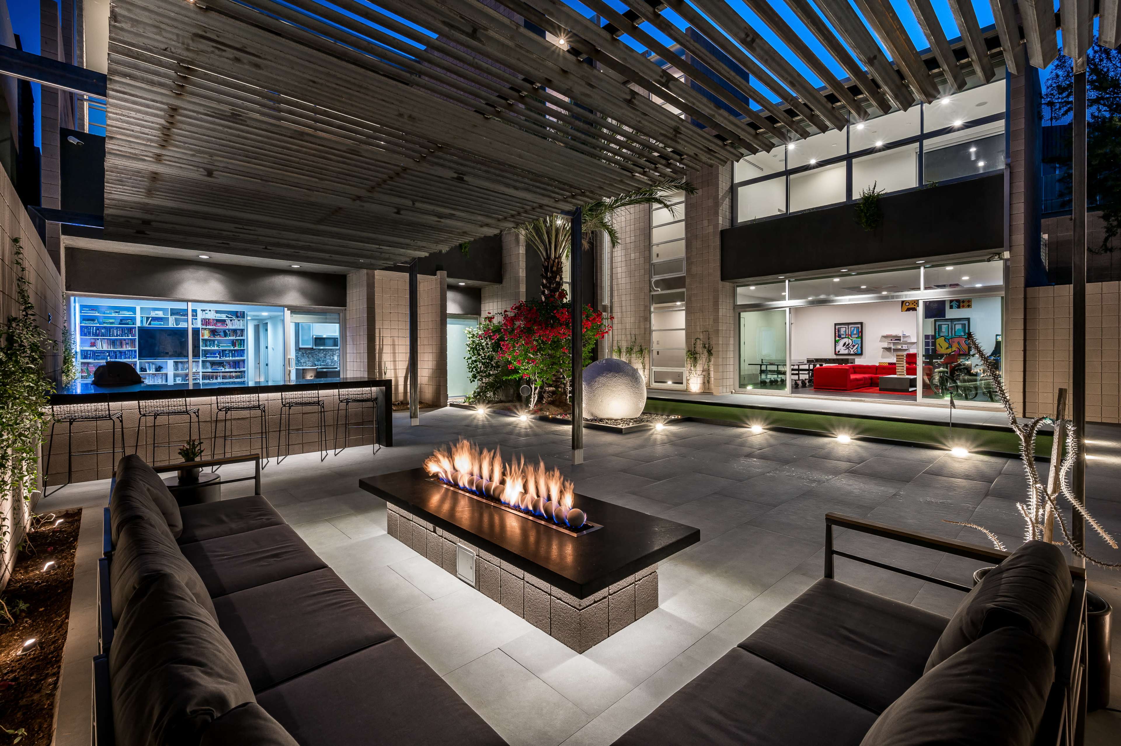 A modern outdoor living space featuring a fire pit surrounded by seating, illuminated at night with a view of a glass-walled interior space.