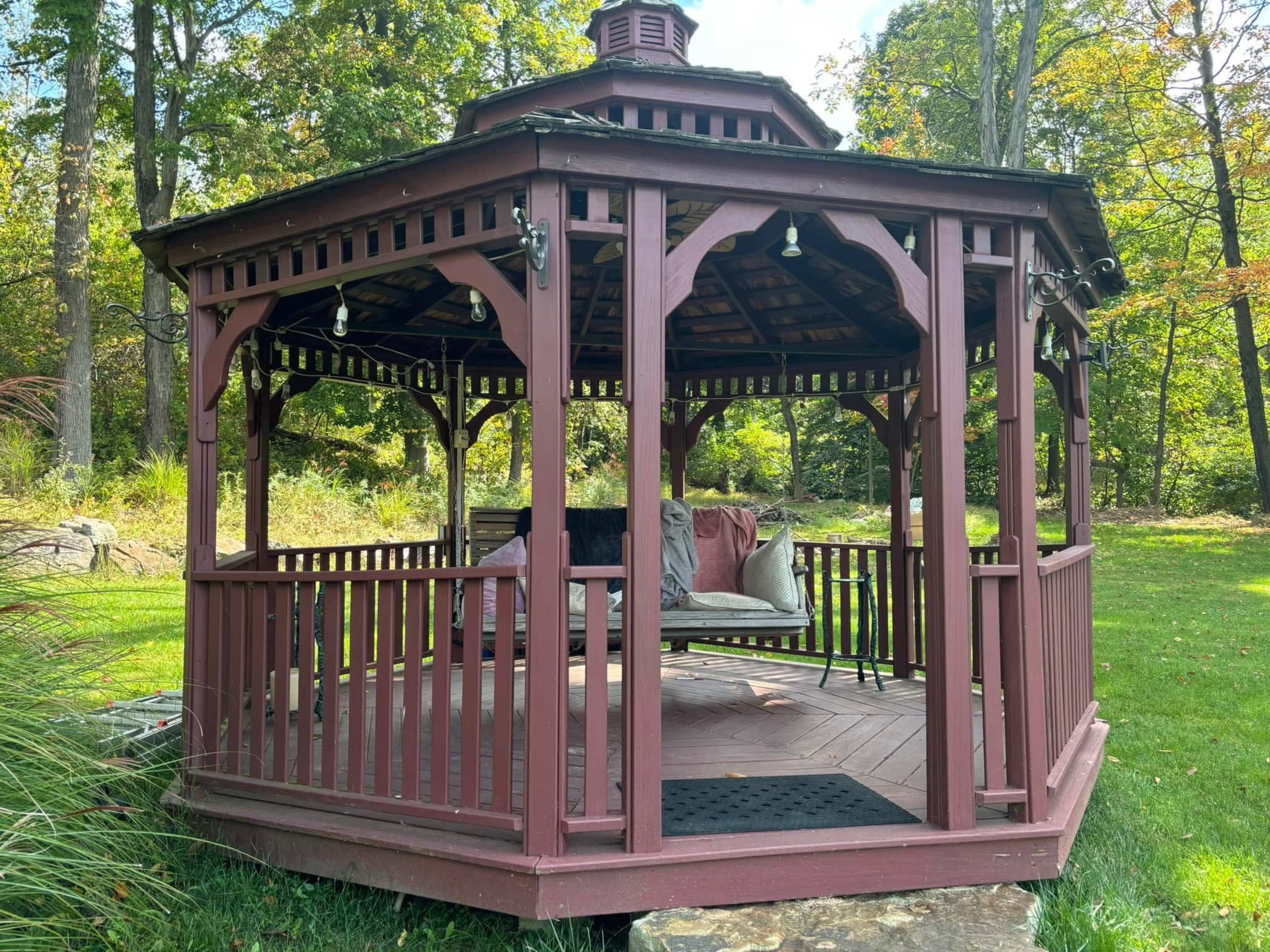 A wooden gazebo with a swing and a lantern is set in a grassy area surrounded by trees.