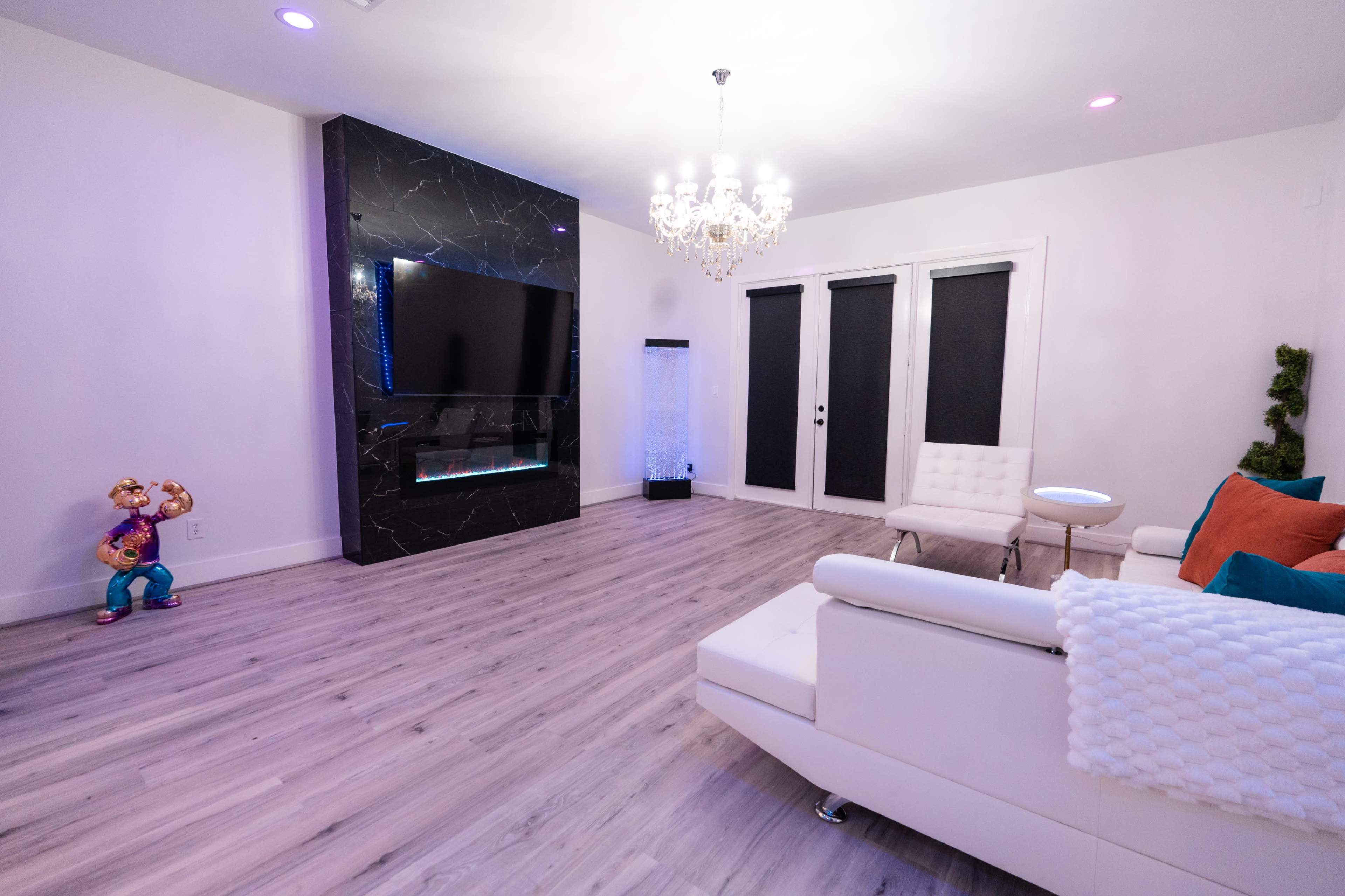 The image shows a modern living room featuring a white sectional sofa, a sleek black accent wall with a mounted television, a chandelier, and decorative elements including a brightly colored figure in the corner.