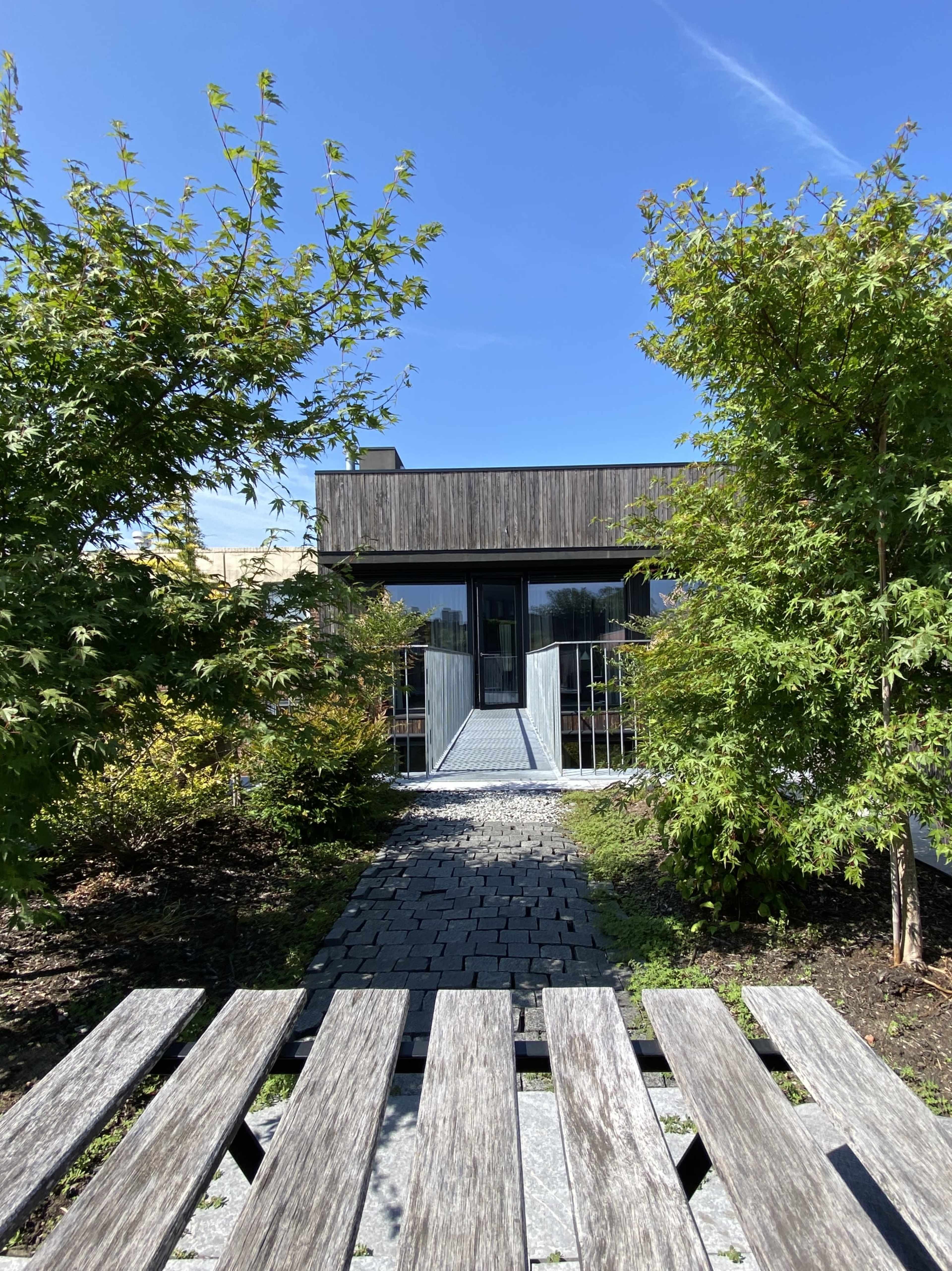 A modern building with a wooden facade is approached via a stone pathway surrounded by shrubs and trees.