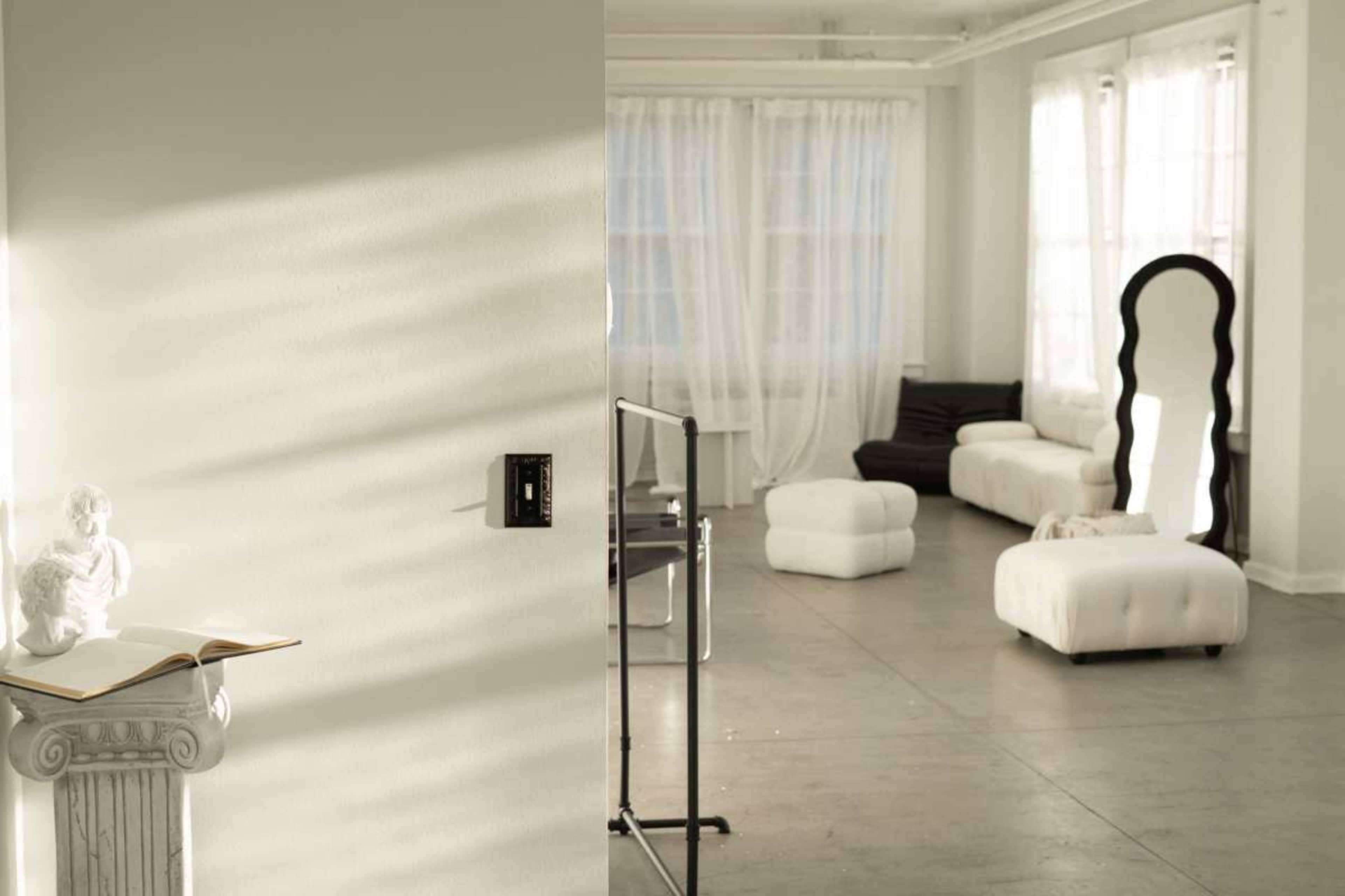 The image shows a bright, minimalist room with large windows, featuring white furnishings, a sculpture on a pedestal, and a mirror reflecting the space.