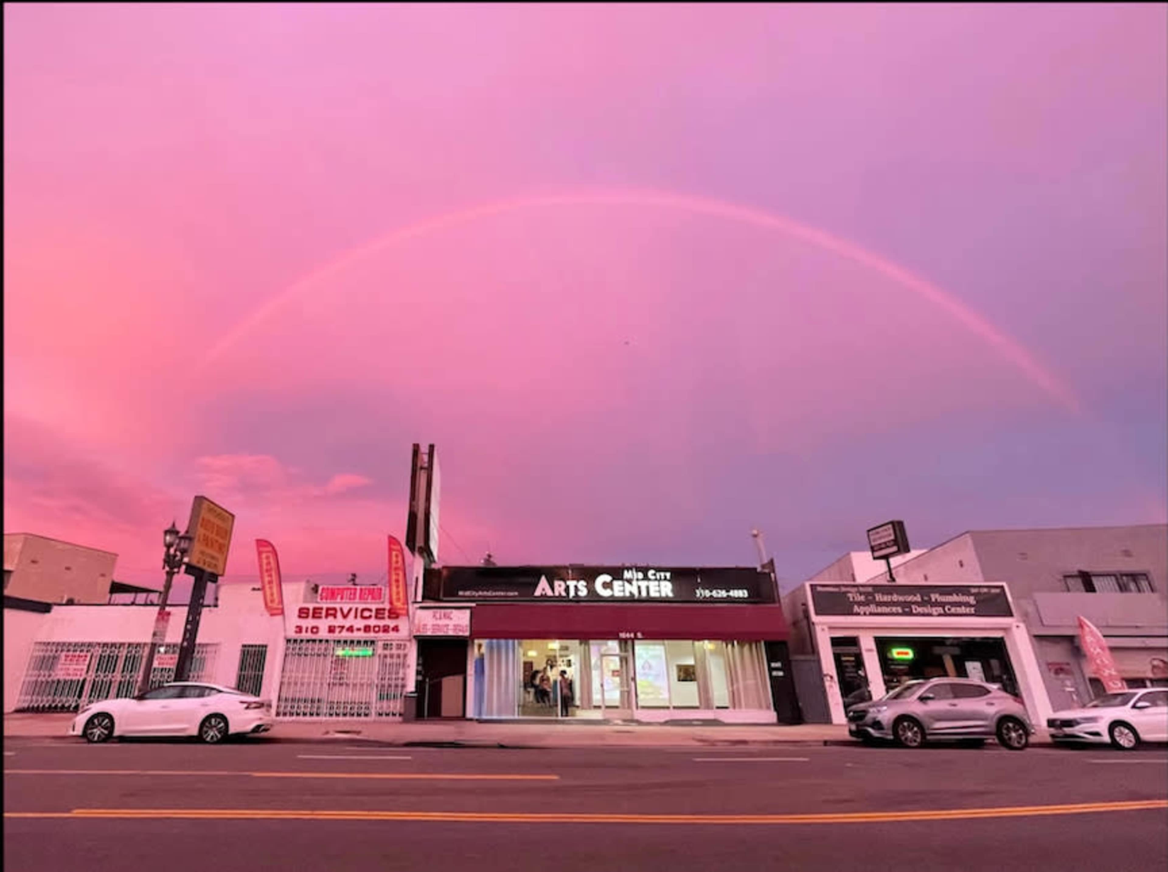 A vibrant rainbow arches over an arts center located on a busy street at dusk.