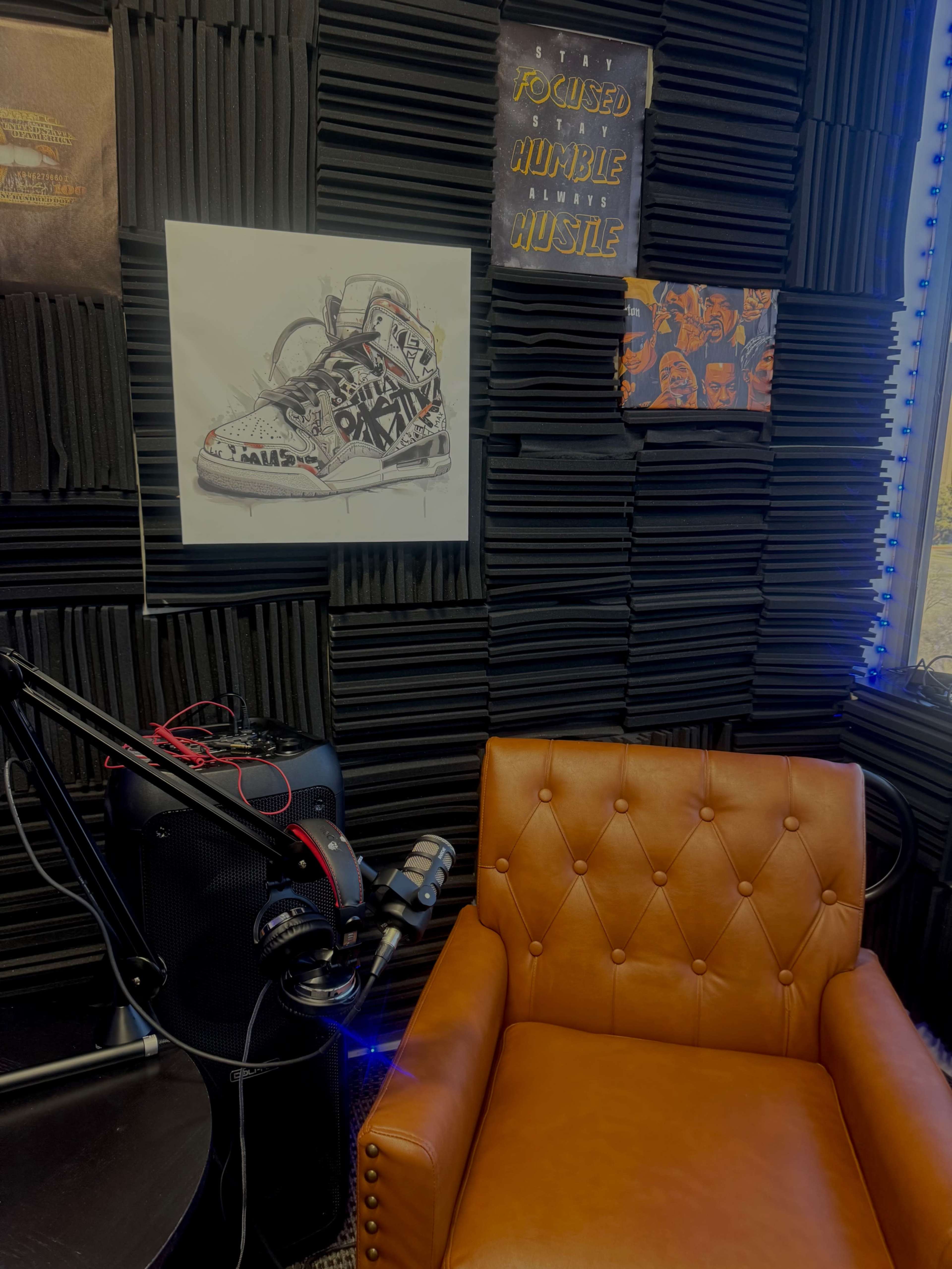 The image features a cozy brown leather armchair next to a microphone and speaker setup against a backdrop of black acoustic panels and vibrant artwork.