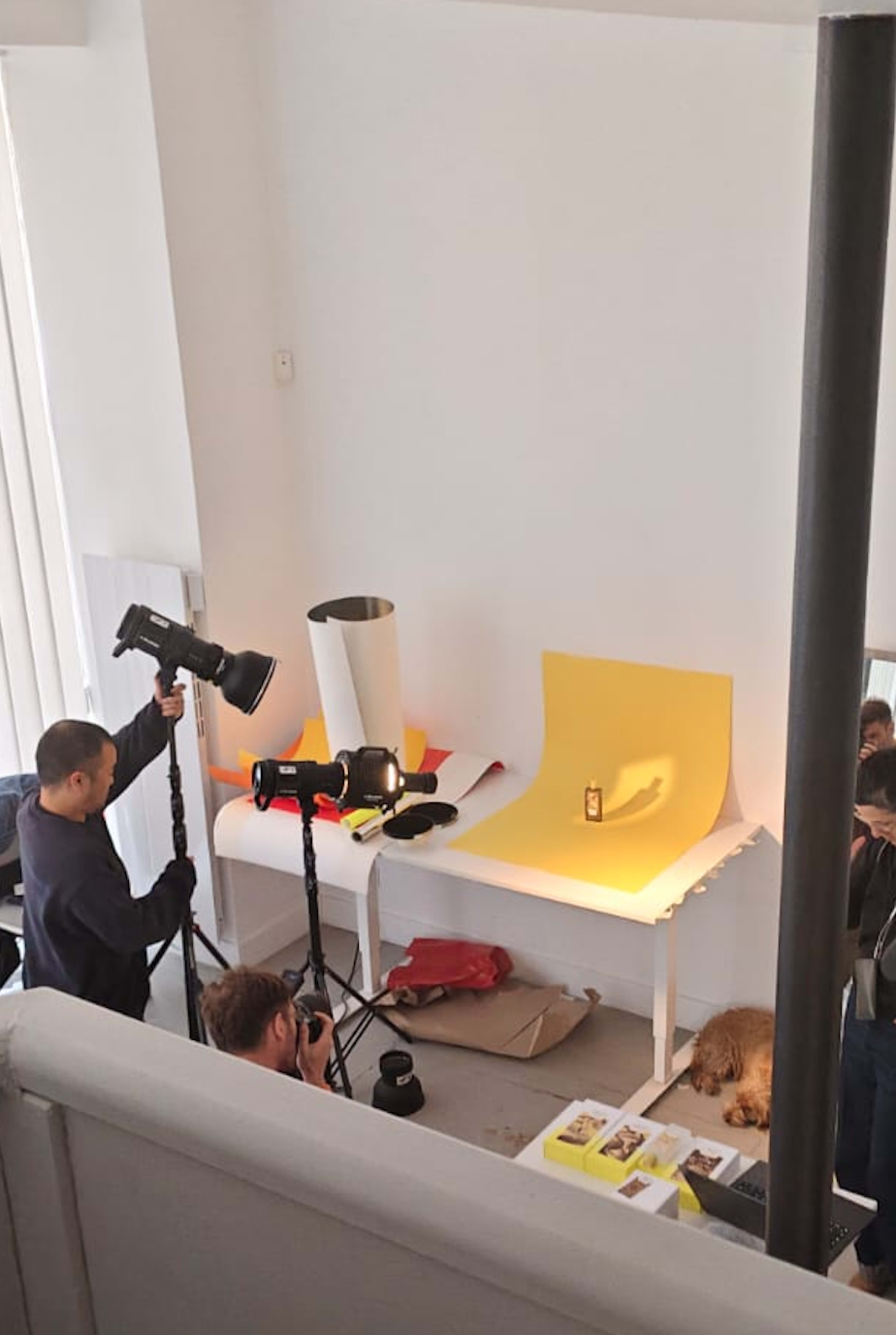 A photography setup features two photographers capturing an object on a table with a yellow backdrop in a bright, minimally furnished room.
