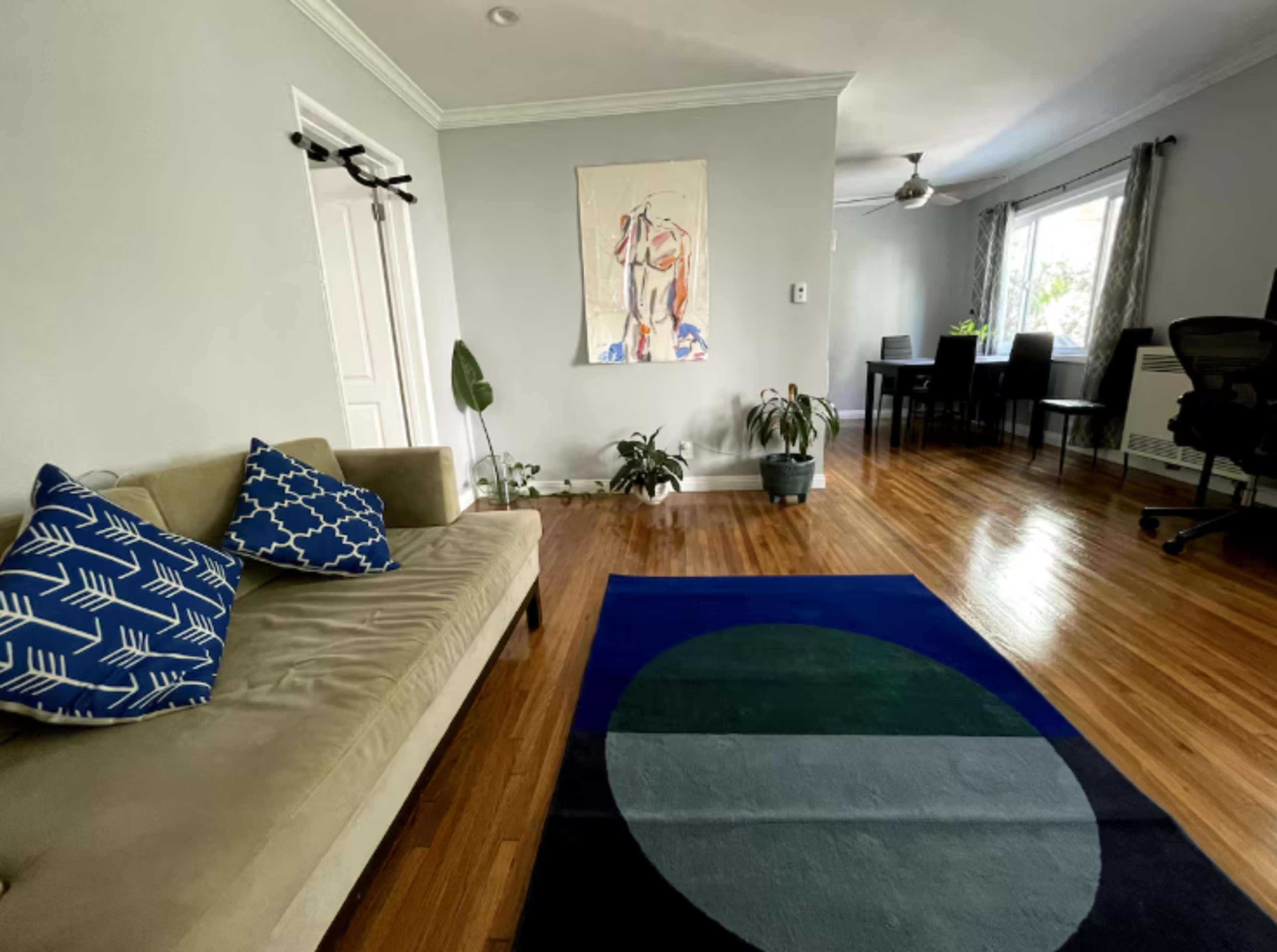 A living room features a light gray wall, a beige sofa with decorative pillows, plants, a colorful area rug, and a dining area with a table and chairs.