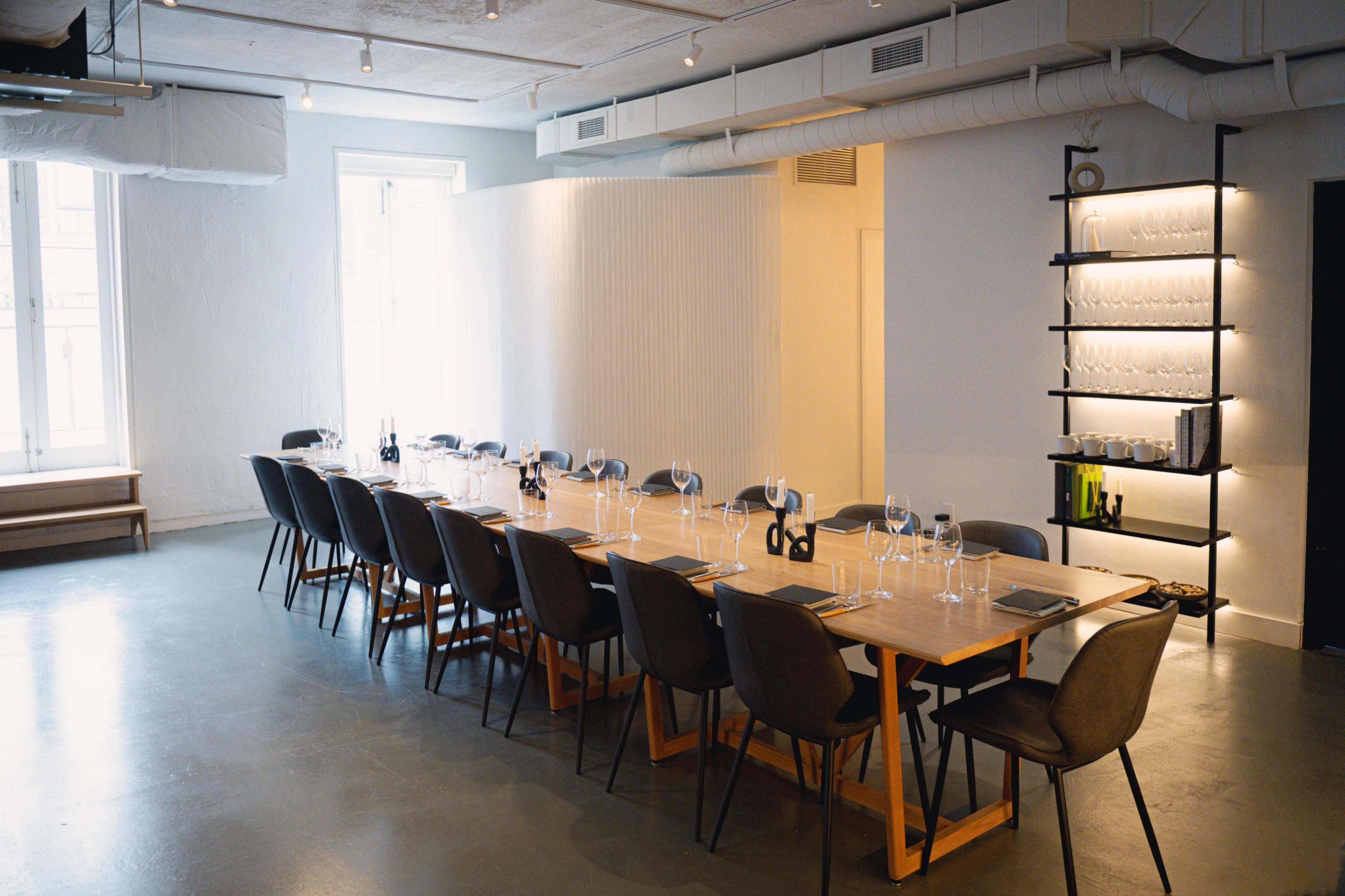 A long wooden dining table is set with glasses and tableware in a modern, minimalist room featuring large windows and a shelving unit.
