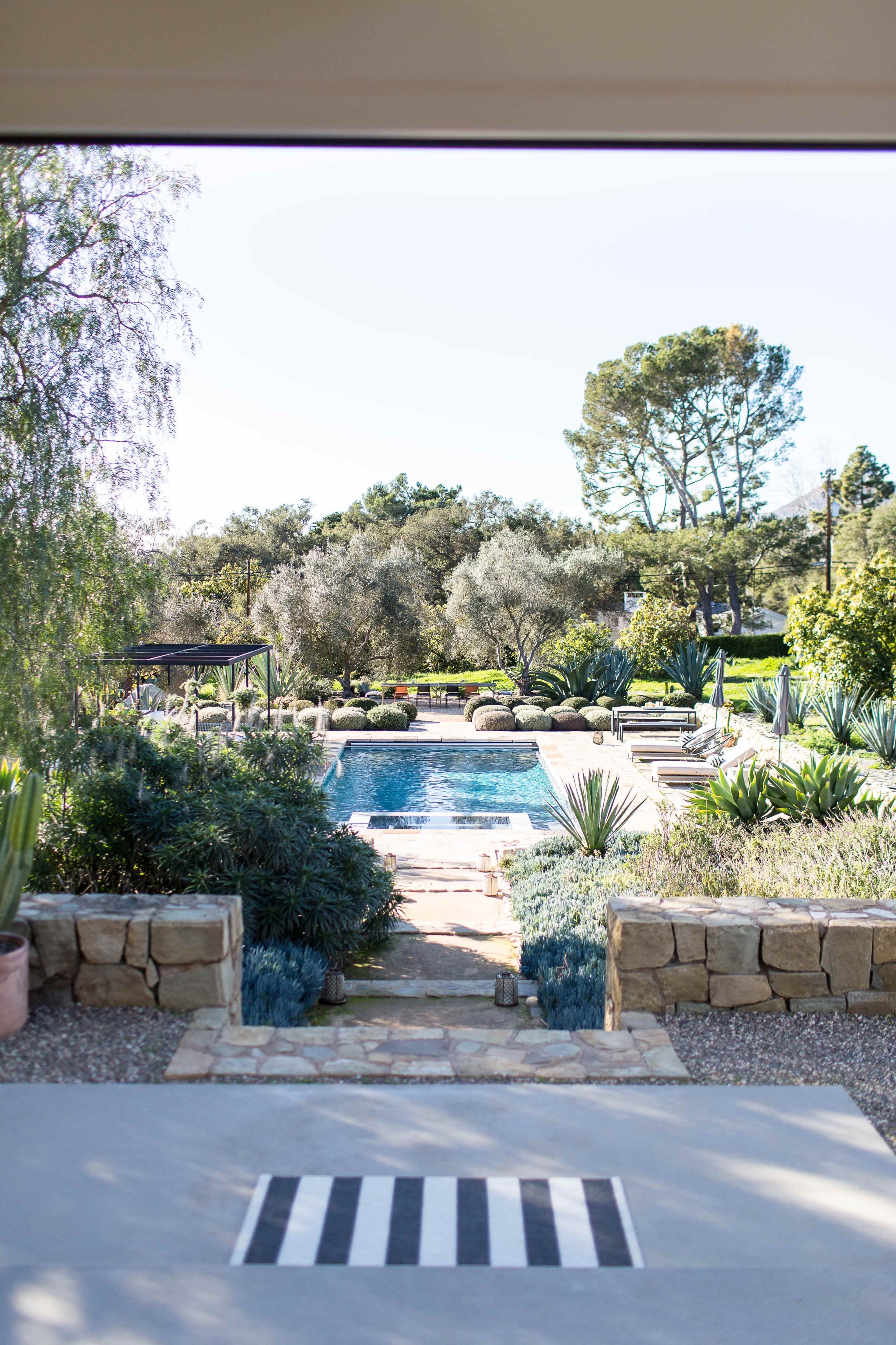 A stone path leads to a rectangular swimming pool surrounded by landscaping that includes various plants and trees.